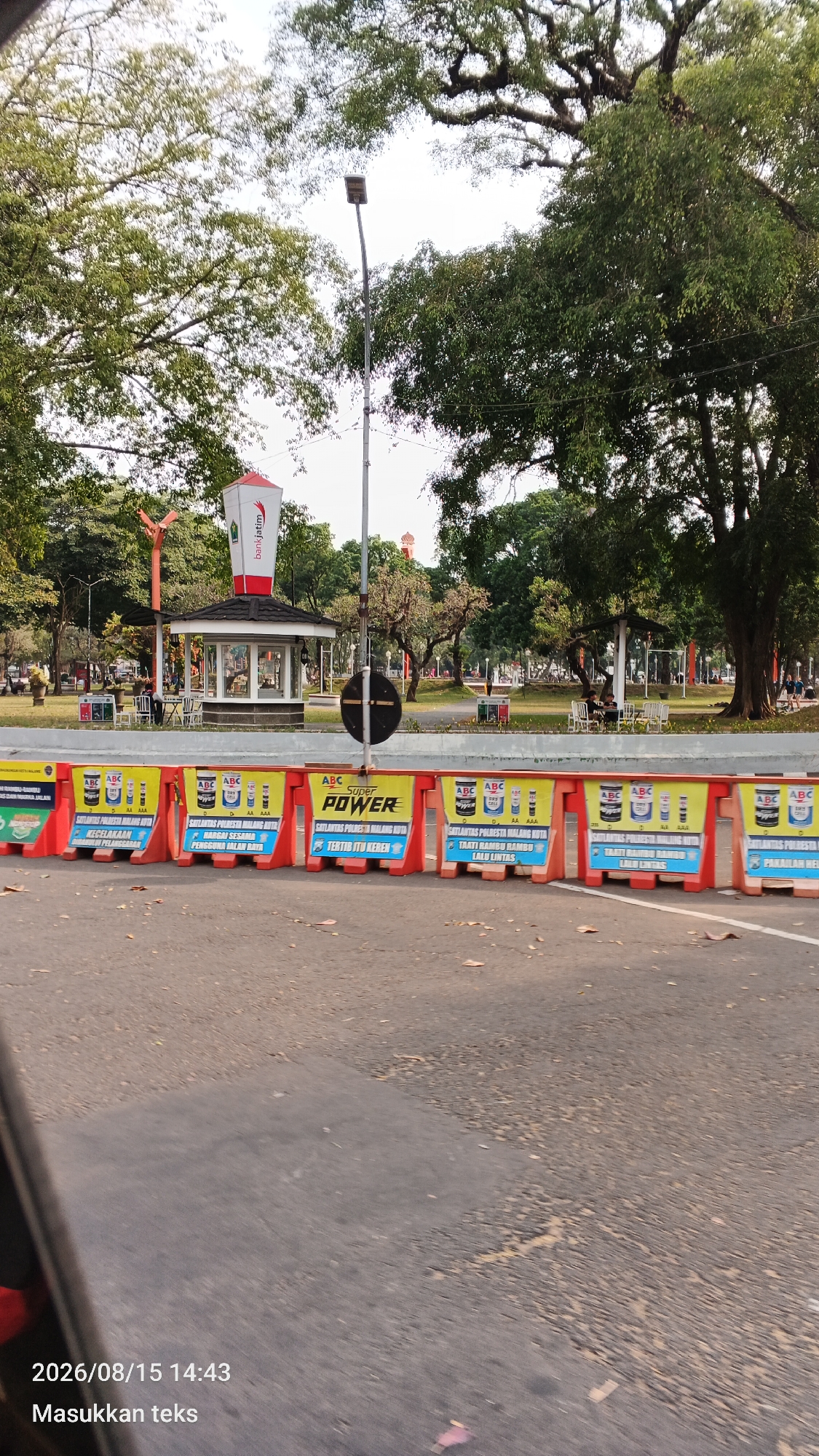 The image depicts a street scene in Malang, Indonesia, with a row of orange traffic barriers in the foreground. These barriers are decorated with advertisements for "ABC Super Power" paint, featuring images of paint cans and text in Indonesian, including slogans related to safety and quality. Beyond the barriers, a park-like area is visible, with trees, grass, and several small structures. One structure resembles a kiosk with a prominent red and white banner featuring the "bankjatim" logo. A lamppost stands near the kiosk. In the background, more trees and a building with a distinct architectural feature, possibly a dome or spire, can be seen. The overall impression is of a public space in a city, possibly a park or promenade, with traffic management in place. The bottom left corner of the image shows a timestamp: "2026/08/15 14:43" and the text "Masukkan teks", which translates to "Enter text".