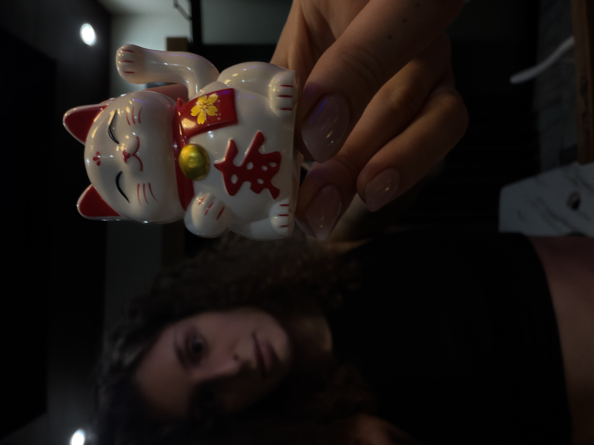 A vertically oriented, close-up photograph features a hand holding a white Maneki-neko figurine over a dark, reflective surface, which shows a blurred reflection of a person.
The upper half of the image is dominated by a fair-skinned hand with manicured nails, painted a light nude color. The hand is gently gripping a ceramic Maneki-neko, or Japanese beckoning cat. The figurine is primarily white, with red-tipped ears, a red collar, and red markings or text on its belly. A golden bell hangs from its neck, and a yellow flower design is visible on a red sash around its chest. Stylized red characters, likely "福" (fuku) for good fortune, are discernible on its stomach. The cat has a smiling expression with closed eyes and one paw raised.
Below the Maneki-neko, reflected on the dark, glossy surface, is the blurred image of a person's head and upper torso. The person has dark, curly hair and is looking forward with a neutral expression, their lips slightly parted. The background above and behind the hand is dark and out of focus, with a few small, bright, blurry light sources suggesting an indoor setting with dim artificial lighting, possibly at night. While the Maneki-neko is Japanese, it's a common decorative item globally and does not contradict the North Kuta, Indonesia location context.