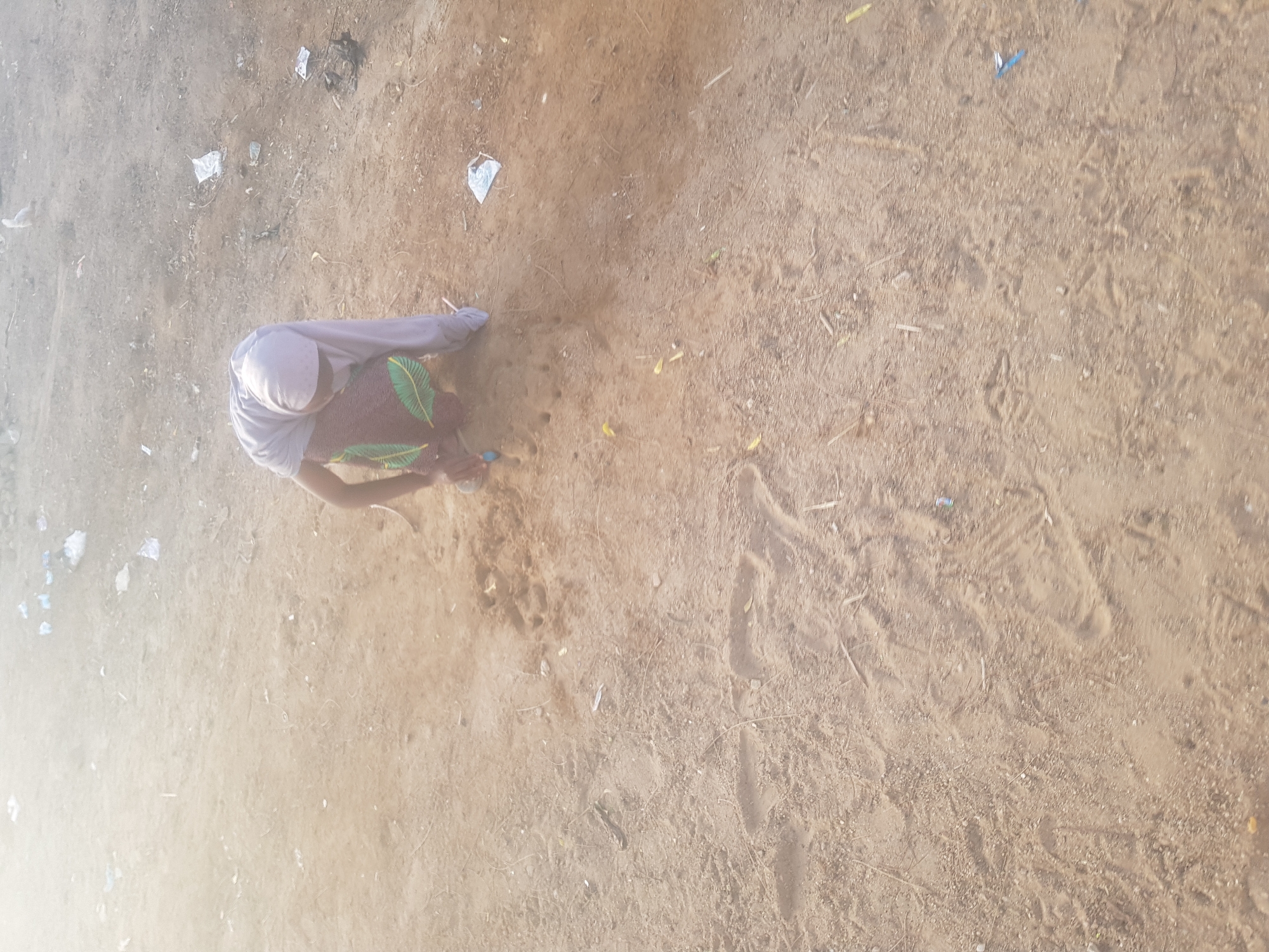 The image shows an individual in Dabawa, Nigeria, squatting on a sandy, dirt-covered surface. The person is wearing a light-colored head covering, a patterned garment, and long sleeves. Debris, including small pieces of paper and other unidentified objects, are scattered around the surface. The environment appears to be an outdoor, possibly undeveloped or unpaved, area within the city. The individual appears to be interacting with something on the ground, but the specific action or object is not clearly discernible due to the angle and distance of the shot.