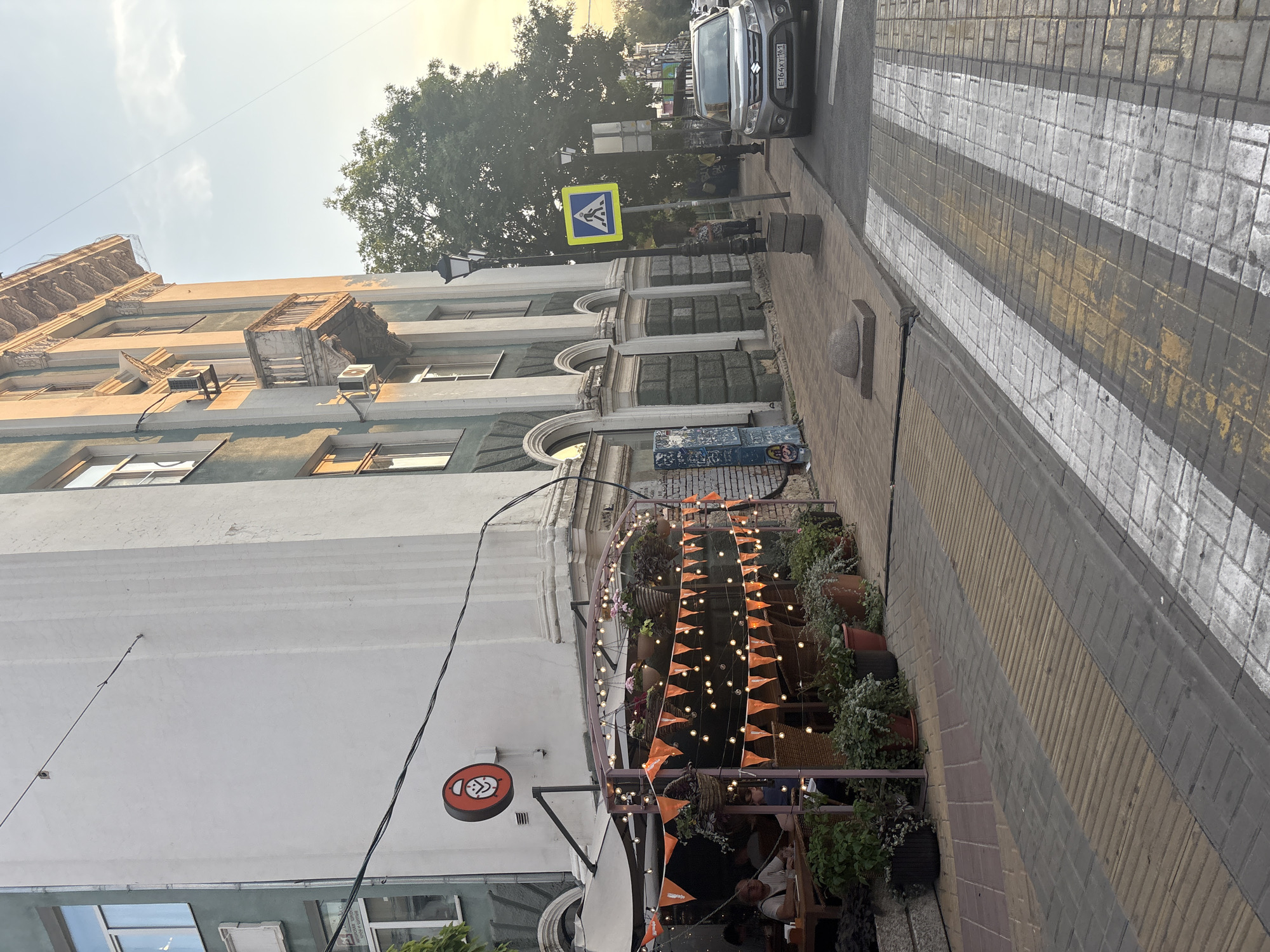 The image shows a street scene in Rostov-on-Don, Russia. A building with an ornate facade is on the left, with a cafe or restaurant area set up on the sidewalk in front of it. The cafe has a covered seating area decorated with orange pennant flags and string lights. Several potted plants are arranged around the outdoor seating.
To the right of the cafe, a sidewalk runs alongside the building, with a street beyond. A pedestrian crossing with white and yellow stripes is visible on the street. A grey Suzuki car is parked near the crossing. A yellow pedestrian crossing sign with a blue symbol of a person walking is mounted on a pole. A lamppost stands on the sidewalk. The sky is clear with a hint of sunset colors. People are seated at tables in the cafe.