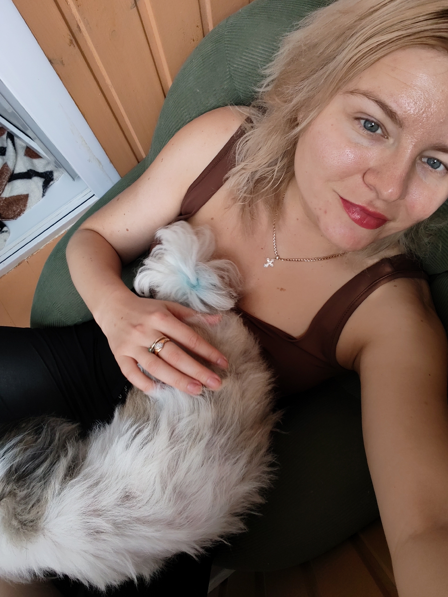A woman with blonde hair and blue eyes, wearing a brown top and black leggings, is holding a small, fluffy white dog with a hint of blue on its fur. She is wearing a gold necklace with a cross pendant and a ring with diamonds. The woman is sitting on a green chair, and behind her is a wall with horizontal wooden planks. To the left, a window reveals a patterned blanket. The scene appears to be indoors, possibly on a porch or balcony, given the wooden wall and proximity to a window. The overall impression is a candid, relaxed moment.