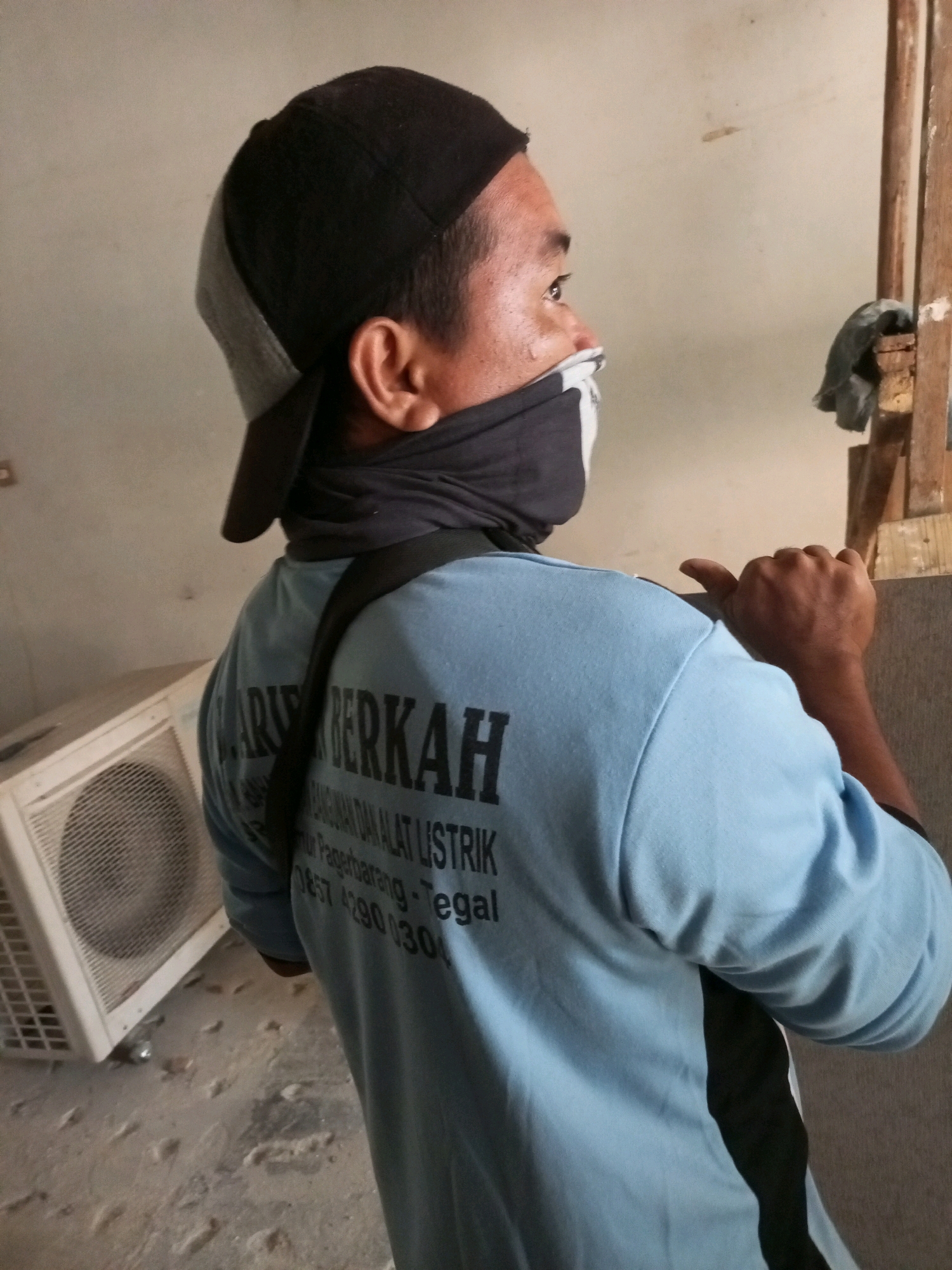 The image shows a man from the back, wearing a blue t-shirt with black lettering, a black baseball cap, and a black face covering. The t-shirt has text that appears to be in Indonesian, including "BERKAH" and "LISTRIK," suggesting it might be related to an electrical service or business. He is wearing a black strap across his chest, and his right hand is raised in a thumbs-up gesture. In the background, to the left, is a white air conditioner unit. The environment appears to be indoors, with a concrete floor and a light-colored wall. The overall lighting suggests an interior space with ambient light.
