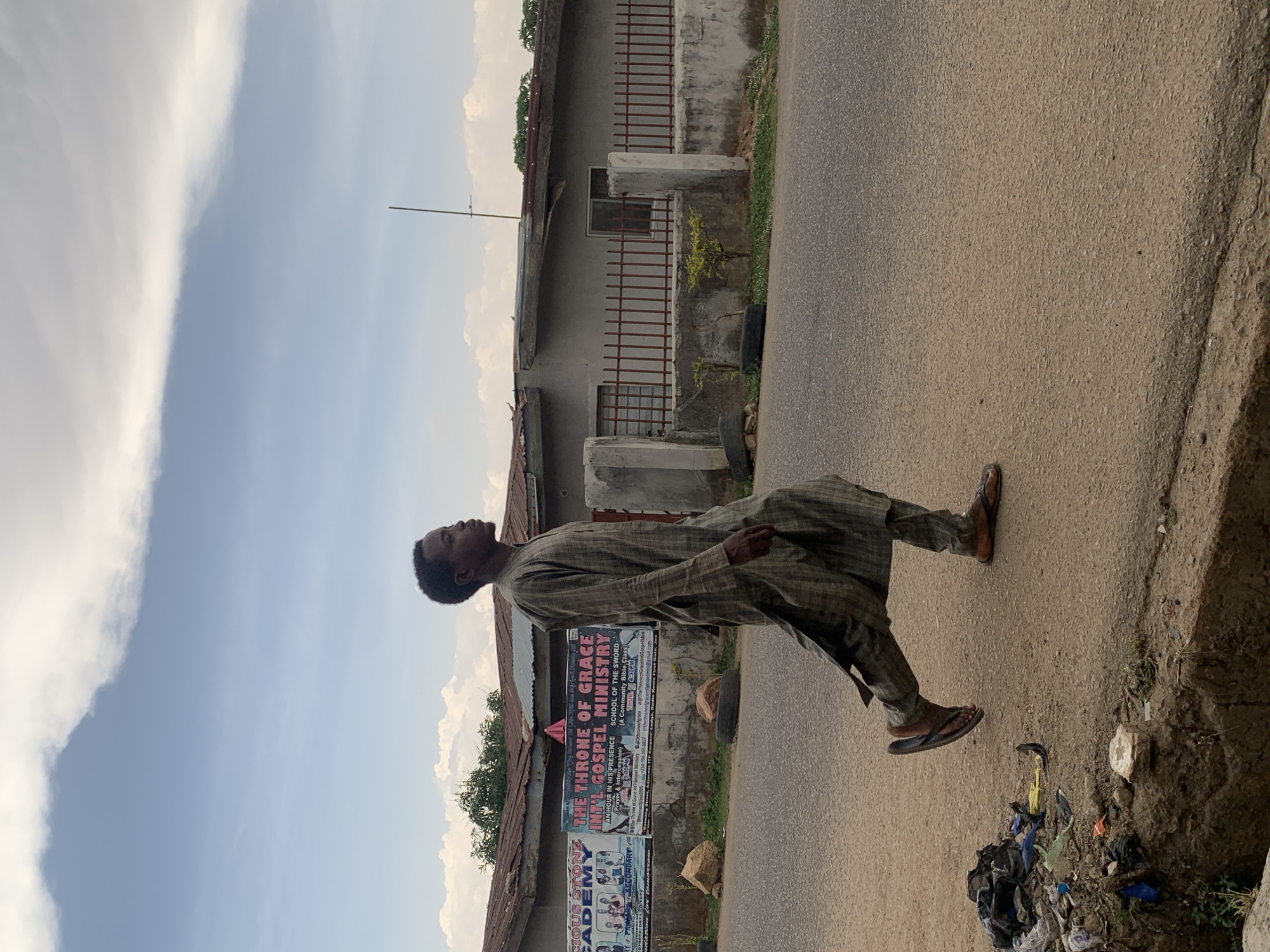 A man stands on a paved road in Gura Top, Nigeria. He wears sandals and a long, plaid garment. Behind him are buildings, including one with a corrugated roof, and posters advertising "The Throne of Grace Int'l Gospel Ministry" and "Precious Stone Academy". Trash is visible at the edge of the road. The sky is visible above the buildings.