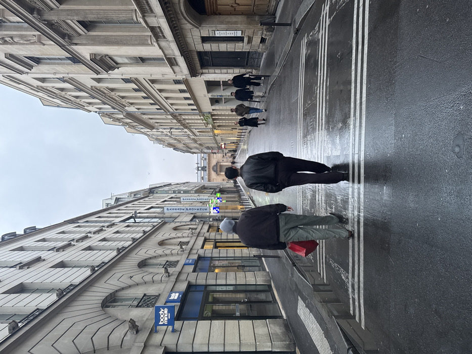 A rainy Parisian street features pedestrians on a slick, marked roadway. The street is lined with tall, ornate buildings, with signage visible above a business named "body minute". Several people walk in the distance, appearing smaller due to perspective. The sky is overcast. The scene conveys a sense of urban life on a wet day in a European city.