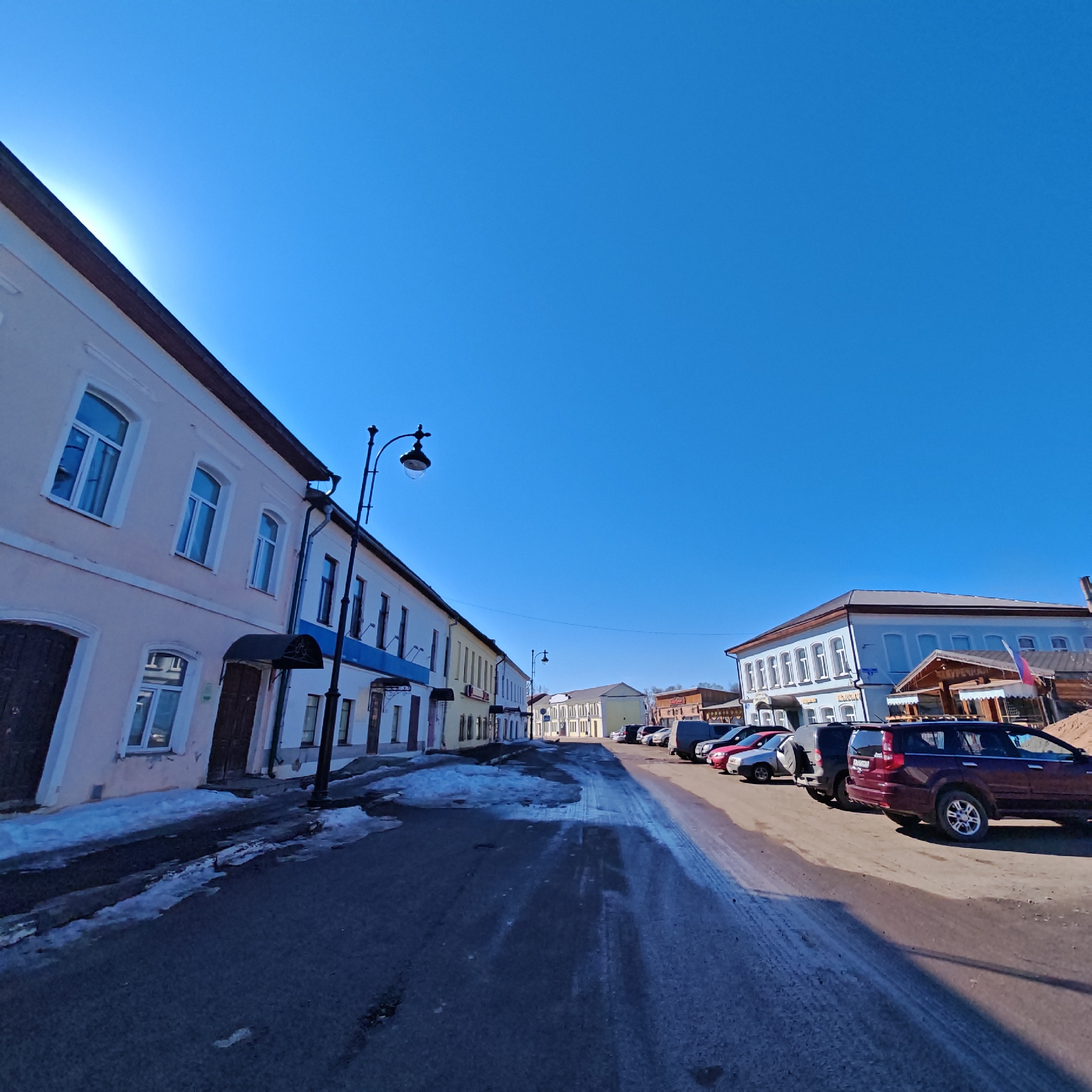 The image shows a street view in what appears to be a historic district of Rostov, Russia. The sky is clear and blue. The street surface is dark with patches of melting snow and ice. Buildings line both sides of the street; the left side features a multi-story building with a peach facade, arched windows, and dark wooden doors, followed by a cream-colored building with a blue awning. On the right side of the street are parked cars of various colors and sizes. The building furthest down on the right has a gray roof, light blue facade and wooden additions. A lamp post is visible near the left side of the street. There are no people immediately visible.