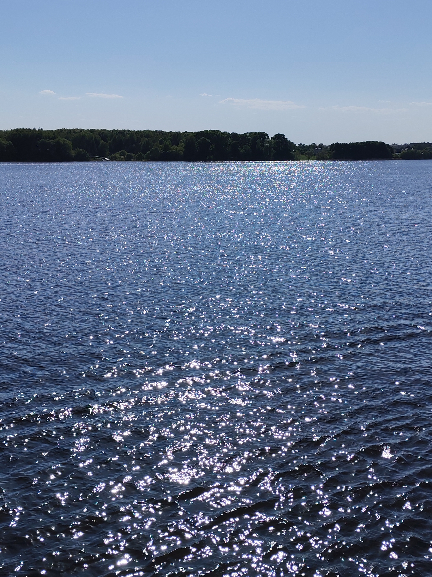 A sunlit body of water stretches toward a treeline in the distance, under a clear blue sky. The water's surface is heavily textured with small waves reflecting sunlight, creating a sparkling effect. The treeline is a solid mass of dark green, with a few hints of structures or clearings visible within it. The overall scene appears to be a natural environment, possibly a lake or river. The location is Kalyazin, Russia.