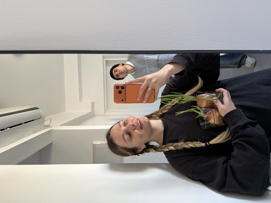 A young adult female is depicted in a mirror selfie, holding an orange-cased smartphone in her right hand to capture the reflection. She has light brown hair styled in two long braids, wears a dark long-sleeved shirt, and a silver ring on her left ring finger. Her expression is a slight smile, and her gaze is directed towards the phone screen. In her left arm, she holds two clear glass tumblers, each containing a brown onion bulb submerged in water with prominent green shoots growing upwards.
Reflected in the background, partially behind and to the right of the female, is a young adult male. He wears a black beanie, a grey crew-neck sweatshirt over a white collared shirt, light blue jeans, and white Crocs. He is looking towards the camera reflection with pursed lips in a playful pose.
The environment is an interior residential space with white walls and a white ceiling. A white split-system air conditioning unit is mounted on the upper wall/ceiling in the reflection. A white door and door frame are visible further in the background. The specific location within the city of Tel-Aviv, Israel, cannot be determined from the visual information provided in the image.