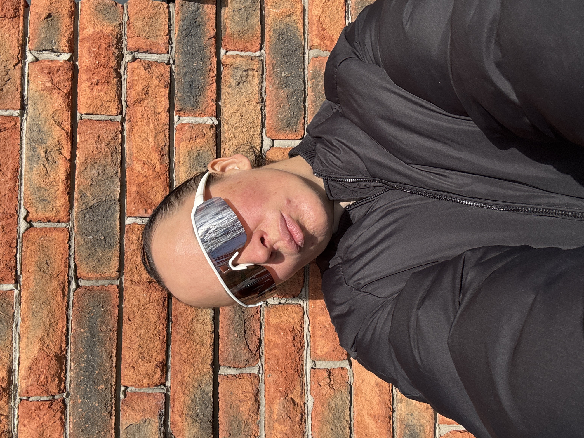 An outdoor portrait, rotated 90 degrees counter-clockwise, depicts a person in a black puffy jacket and large, white-framed mirrored sunglasses. The person's head is positioned on the left side of the frame, angled slightly upward and looking towards the viewer, with their body extending to the right. They are set against a rustic brick wall, which features rough-textured bricks in shades of orange, red, and dark grey, joined by lighter-colored mortar.
The scene is brightly lit by strong sunlight, which creates prominent reflections on the mirrored lenses of the sunglasses and distinct highlights on the fabric of the jacket. The light source appears to be at a low angle, suggesting either morning or late afternoon. The person's facial expression is neutral, largely obscured by the sunglasses, which reflect a distorted image of the surrounding environment. Their dark hair is visible at the temples. There are no visible logos or text. The overall atmosphere is clear and sunny. The location is Chaikovsky, Russia.