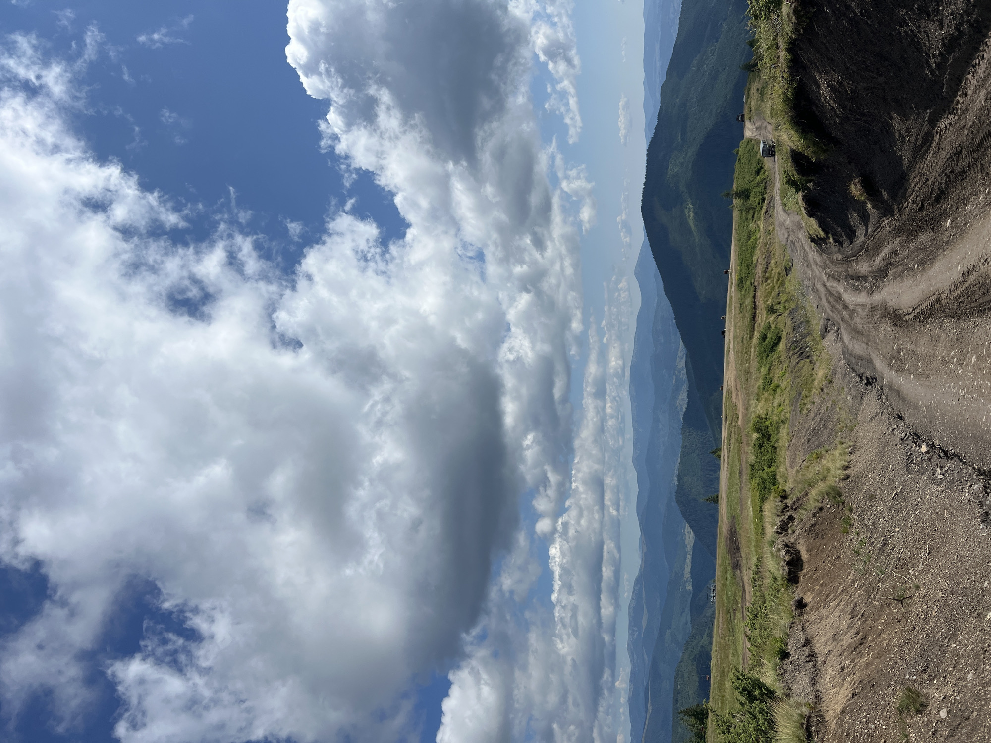 This is a full-frame, eye-level, outdoor, vertical shot of a mountainous landscape in Chorna Tysa, Ukraine. The foreground is dominated by a dirt road with tire tracks, which curves from the bottom right of the frame into the middle distance. The road is bordered by uneven, grassy terrain and some exposed soil and rocks on the right. In the mid-ground, there are rolling green hills with sparse trees and a few off-road vehicles visible on the road and the surrounding fields. The background features a series of layered, dark green mountains receding into the distance, with a hazy, blue-toned sky. The sky itself is partly cloudy, with large, dramatic white and gray clouds interspersed with patches of bright blue sky. The lighting suggests it is daytime, with the sun casting shadows and illuminating the landscape. There are no identifiable people visible in the image.