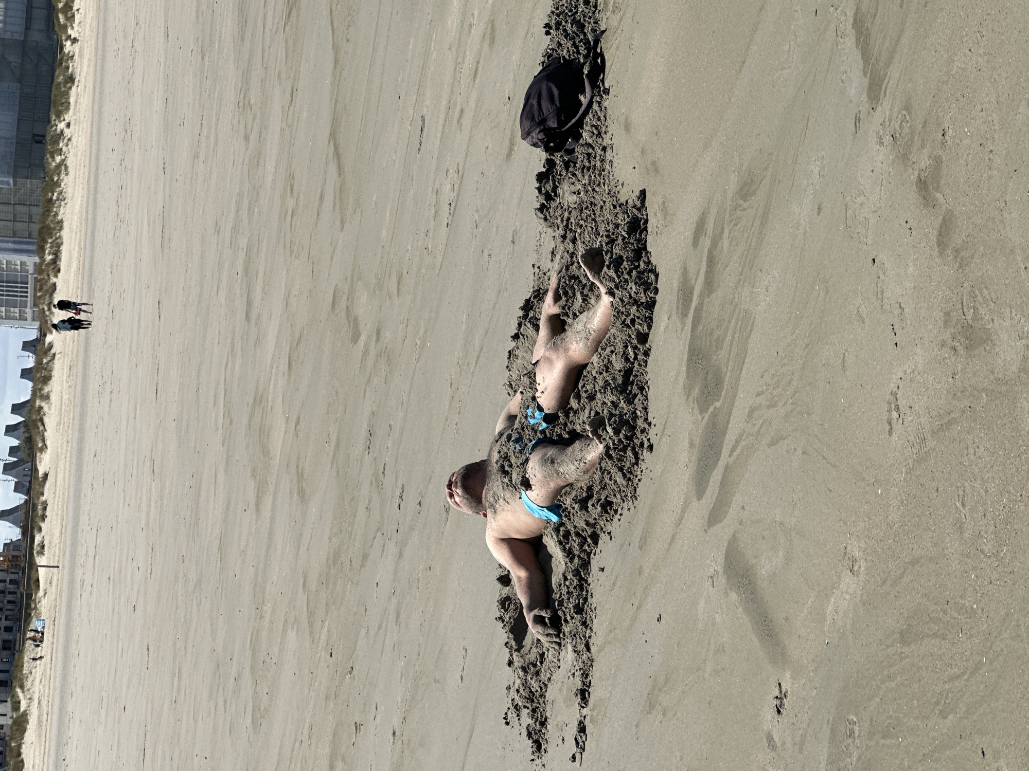 In Dunkirk, France, a man lies buried in the sand on a beach. He is wearing blue swim trunks and has his eyes closed, facing upwards. His arms are outstretched to his sides, and his legs are covered by sand. A black bag rests on the sand to his right. In the background, two people walk along the beach, and behind them, a row of buildings and dunes are visible. The scene is bathed in sunlight, casting shadows on the sand.