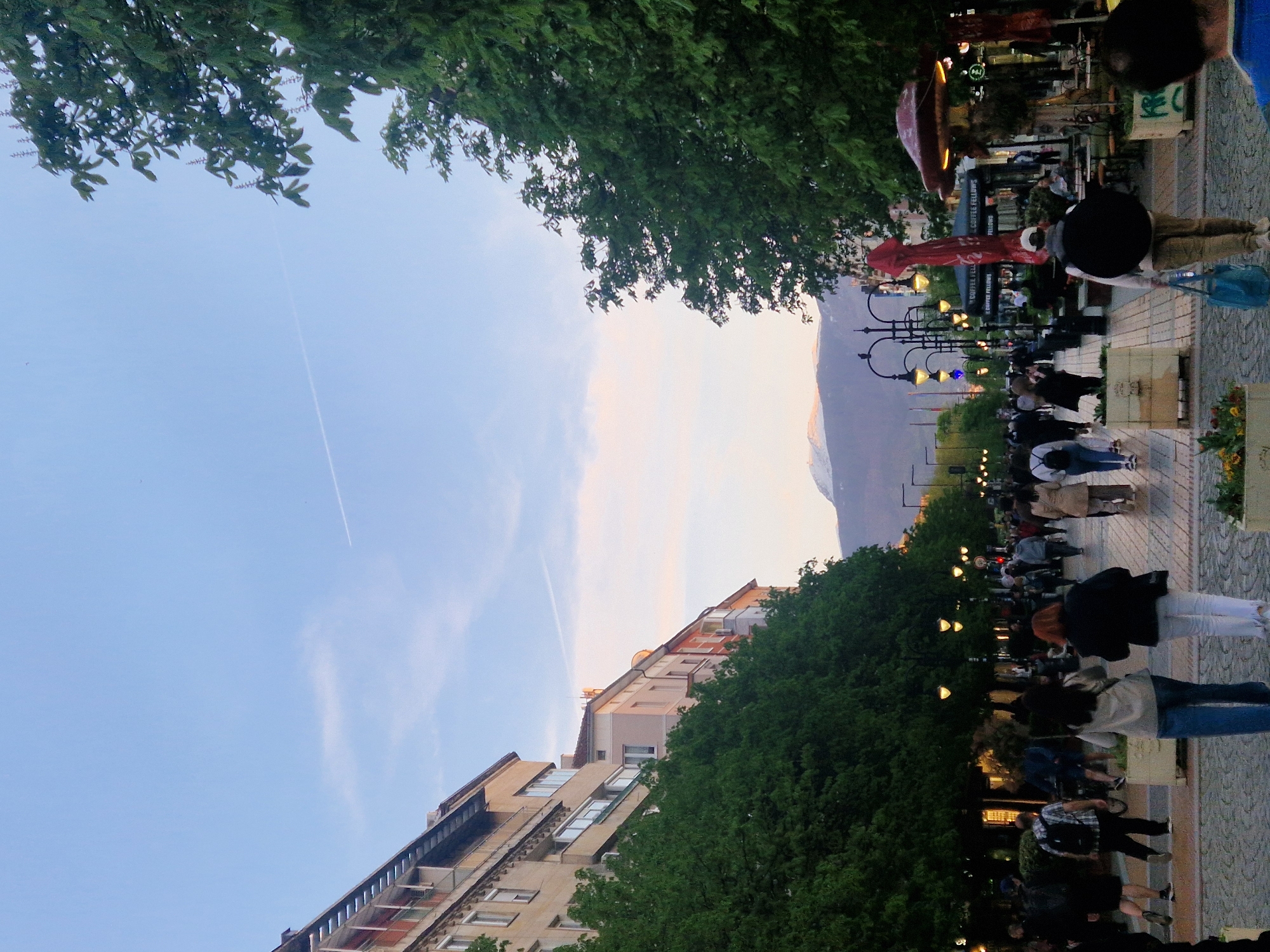 The image depicts a lively pedestrian street scene, likely a promenade, during the late afternoon or early evening. A wide, paved street is bordered by lush green trees and buildings.
Numerous casually dressed people are walking, standing, and possibly sitting at outdoor cafe tables, contributing to a bustling atmosphere. On the left side (when upright), multi-story buildings with balconies and windows are visible. The right side of the street is lined with traditional lampposts, which are already illuminated with warm yellow and cooler blue lights. Red umbrellas and outdoor seating suggest cafes or restaurants along this stretch.
Lush green deciduous trees form a canopy over parts of the street, particularly at the top and bottom edges of the frame. In the distance, a prominent mountain range stands under a gradient sky, transitioning from a clear blue at the top to lighter, possibly pinkish or yellowish hues near the horizon. A faint airplane contrail streaks across the upper sky.
The activity suggests people are enjoying a leisurely evening stroll, socializing, or dining outdoors. The time of day is indicated by the soft, fading natural light combined with the glowing artificial streetlights. The weather appears clear and pleasant. Notable details include the text "COFFEE FELLOWS" visible on a sign and an umbrella, identifying a cafe, and some green graffiti on a pillar. A blue shopping bag is carried by one person.
Location context: Unknown, Unknown