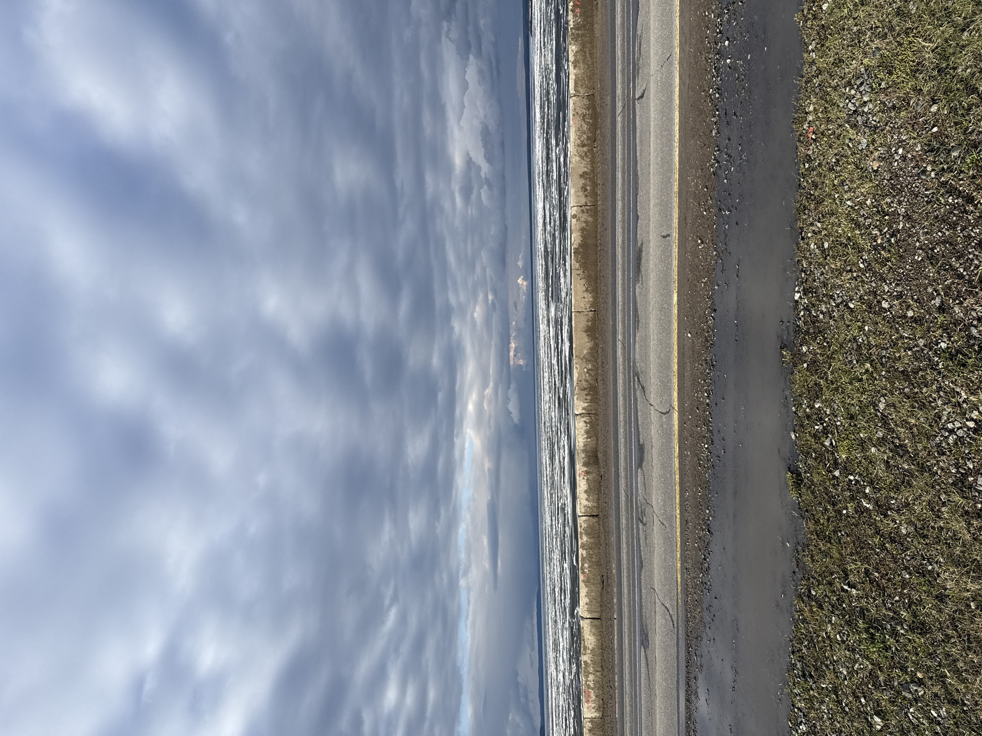 This image, when correctly oriented, displays a landscape featuring a large body of water, a road, and a cloudy sky. The scene is set along what appears to be a vast expanse of water, likely the Votkinsk Reservoir in Chaikovsky, Russia, given the provided context. The water surface is highly reflective, suggesting either ice floes or turbulent water with strong reflections from a bright, overcast sky.
Running parallel to the water in the middle ground is a paved asphalt road. It has a distinct solid yellow line marking its edge, and visible cracks indicate some wear. Between the road and the water, there's a narrow, dark strip of ground, possibly a wet curb or shoulder. On the other side of the road, to the viewer's right, lies a patch of unkempt ground covered with sparse, dormant grass, dark soil, and numerous small stones and pebbles, suggesting a natural, uncultivated area.
The sky, dominating the left portion of the frame, is filled with varying densities of grey and white clouds, indicating an overcast or partly cloudy day. The lighting is bright but diffused, suggesting daytime, possibly during a colder season given the appearance of the landscape and water. There are no people or specific activities visible, creating a static and somewhat serene atmosphere. No discernible text is present in the image.