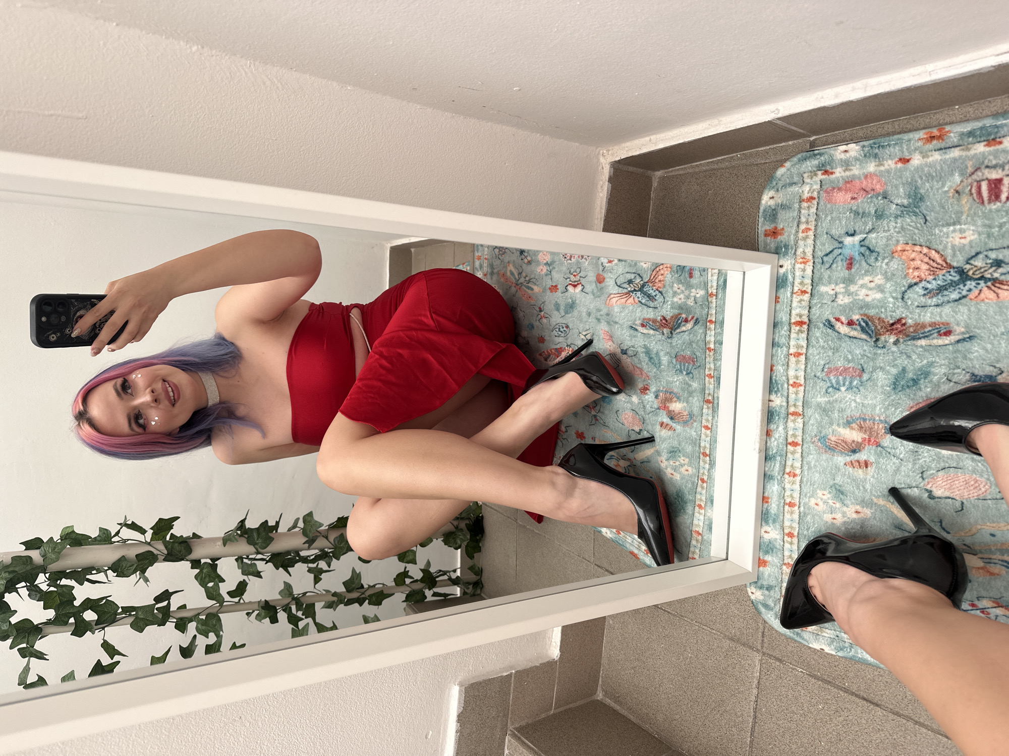 A mirror selfie shows a young woman with purple and pink hair lying on a patterned rug while wearing a red dress and black high heels. She is holding a black smartphone to take the photo. The setting appears to be an indoor space with a white wall and tile flooring, with artificial ivy plants visible in the mirror's reflection. Although the exact location cannot be pinpointed from the image, it is specified to be in the city of Zagreb, Croatia.