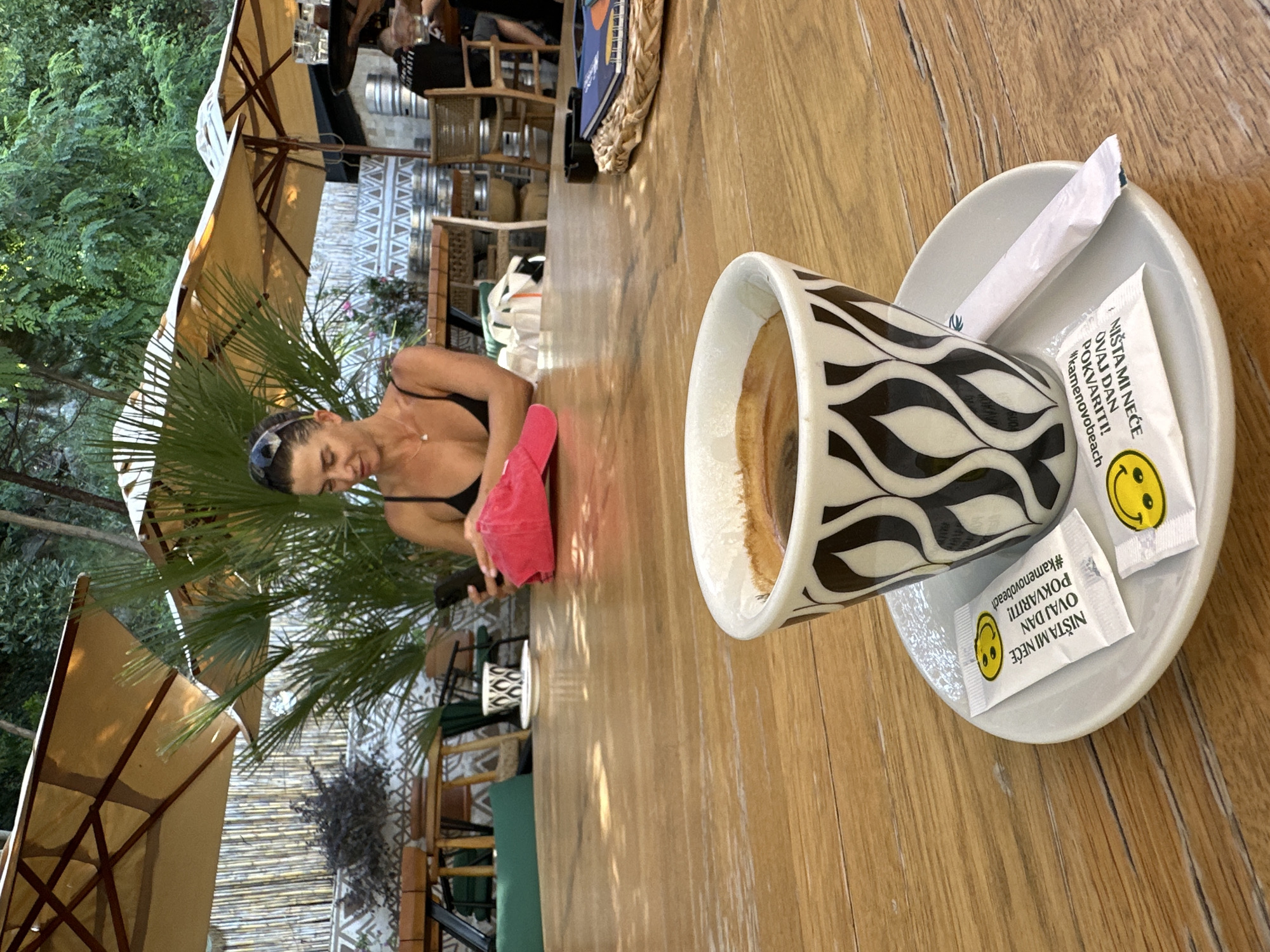 A woman with dark hair pulled back, wearing a black bikini top and sunglasses on her head, is seated at a wooden table outdoors. She is looking down at her phone, which she is holding in her right hand. In her left hand, she holds a bright pink towel or cloth. A coffee cup with a black and white wavy pattern sits on a saucer in the foreground, filled with coffee. Two packets of sugar, one with a smiley face and text in Croatian that translates to "Nothing will spoil this day!", are placed on the saucer next to the cup. Behind the woman, there are large beige umbrellas providing shade, palm fronds, and bamboo fencing, suggesting a tropical or beachside setting. In the background, there are tables, chairs, and people seated, indicating a cafe or restaurant. The lighting suggests it is daytime.