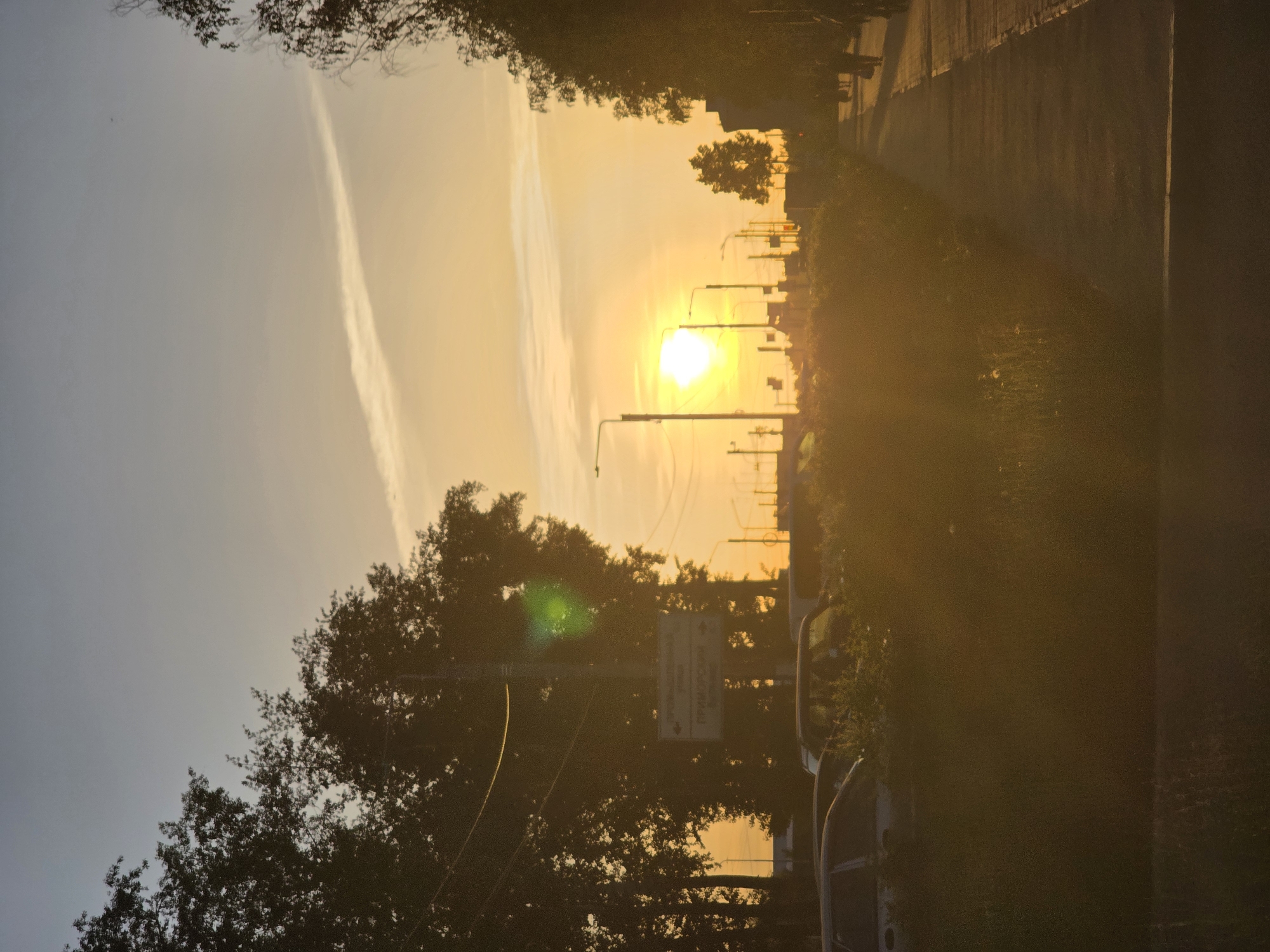 The image captures a sunset in Chaikovsky, Russia. A road stretches into the distance, flanked by trees and vegetation. A contrail streaks across the sky. Streetlights are visible against the setting sun. Parked cars are also seen.