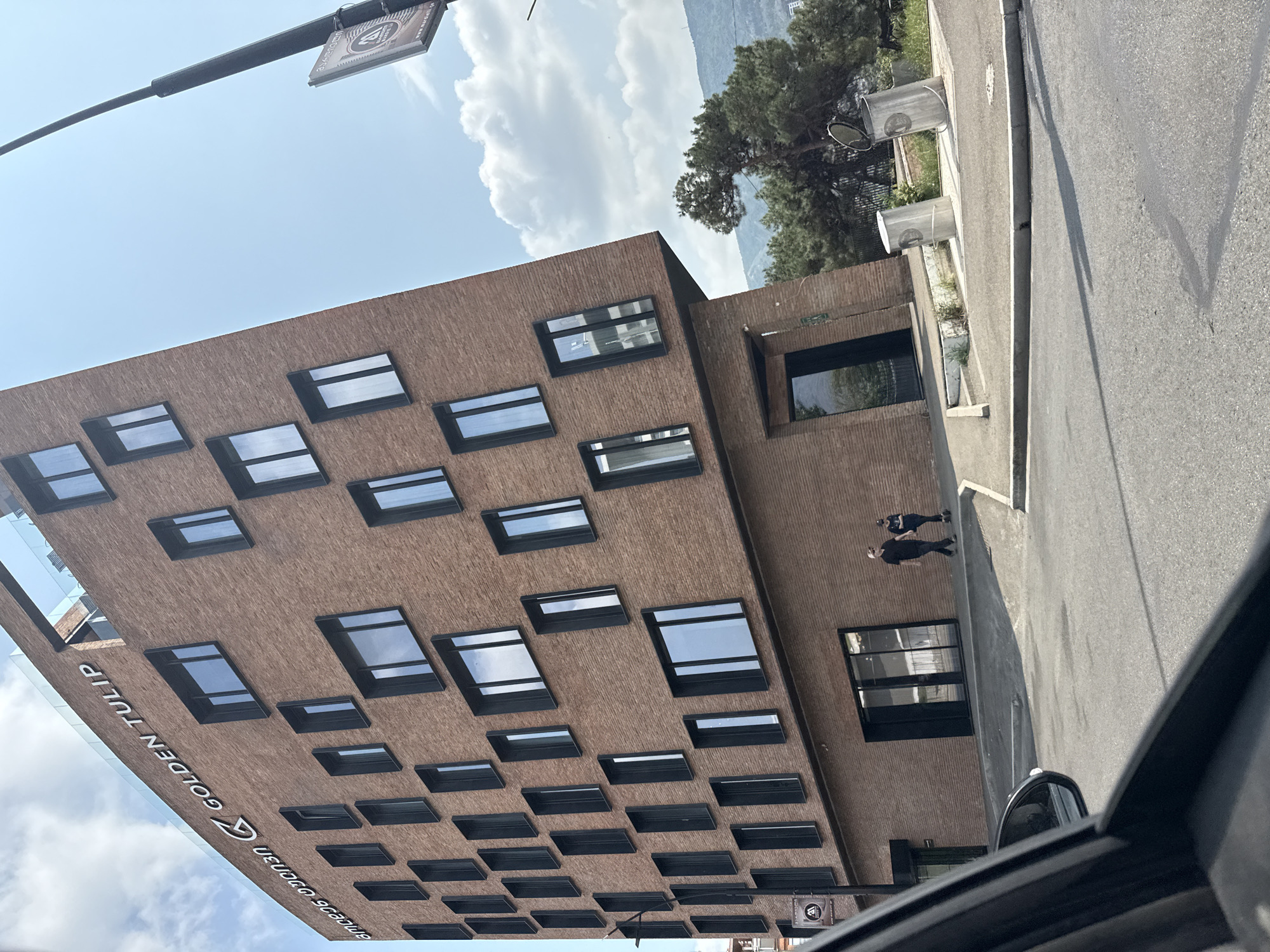 In an urban environment in Tbilisi, Georgia, a modern, multi-story building with a brick facade dominates the frame. The building features a contemporary design with numerous rectangular windows of varying sizes, some of which are darker and appear to be tinted. The words "GOLDEN TULIP" are visible in large white lettering on the upper facade, along with a stylized "G" logo. Adjacent to the building, a street is visible with a sidewalk and two metal trash receptacles. Two individuals are present on the sidewalk: one person, likely a male, wearing a dark t-shirt and light-colored cap, is walking away from the camera, and another person, appearing to be a female, is also walking away from the camera and is on a scooter. The scene is captured from a low angle, possibly from inside a vehicle, as a portion of a car's side mirror and windshield frame are visible in the lower left corner of the image. In the background, lush green trees and a distant hillside are discernible under a clear blue sky with scattered white clouds. A lamppost with a sign attached to it is also visible to the right of the building.