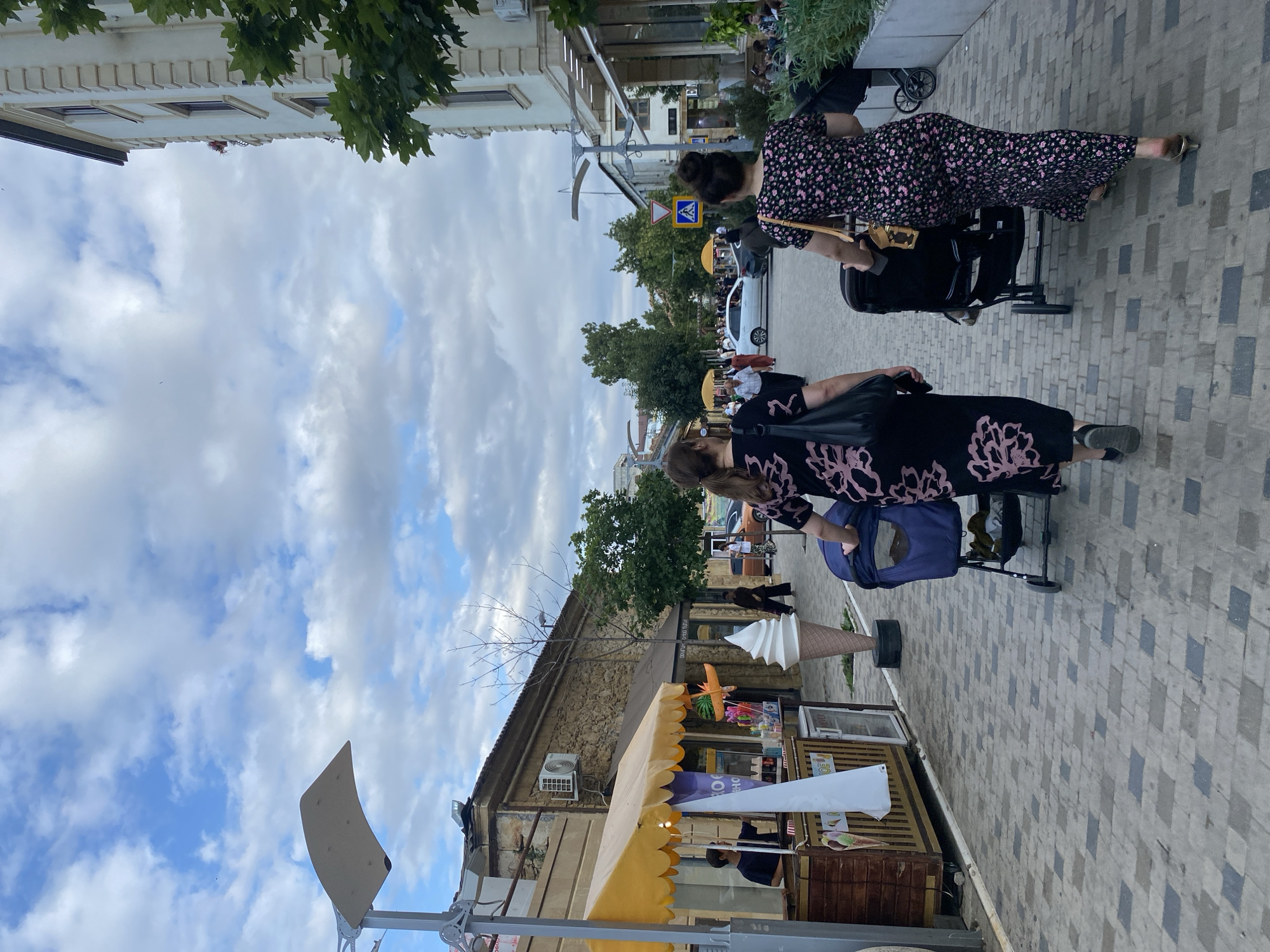 This image captures a street scene in Derbent, Russia. The foreground features two women walking on a patterned brick sidewalk. One woman, in a black dress with a pink floral pattern, pushes a stroller. She is wearing sandals. The other woman, also in a black dress with a pink floral pattern, is partially visible in the foreground, facing away from the camera.
Behind the women, a street lined with buildings and trees unfolds. A yellow awning covers a small shop selling various items, including what appears to be a large decorative ice cream cone and other novelty items. Signs with text are visible on the shop, but are not clearly legible. Further down the street, more buildings, trees, and a white car are visible.
The sky is cloudy with patches of blue, suggesting it is daytime. The overall atmosphere seems casual, with people strolling along the street, possibly enjoying an outing. The architecture hints at a historical or older part of the city. There are no strong indicators of emotion, but the presence of people and shops suggests a lively, public space.