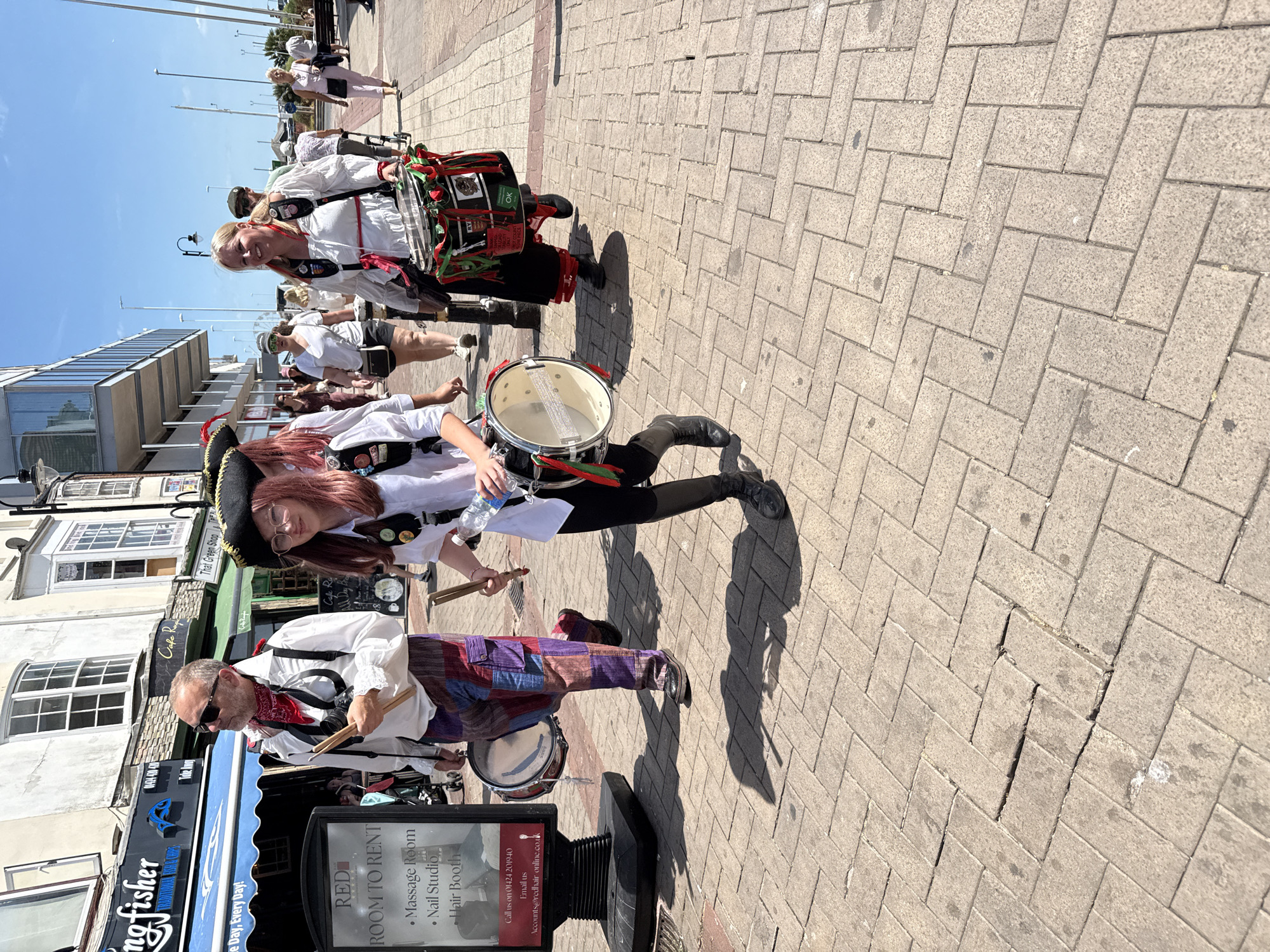 The image shows a street scene in St Leonards, United Kingdom, with several people in historical or theatrical costumes marching and playing drums. In the foreground, a person with pink hair, wearing a black pirate hat, glasses, and a white ruffled shirt, is playing a drum. To their left, a man with sunglasses and a red bandana around his neck, also in a white ruffled shirt, carries drumsticks. Further to the right, a woman in a white ruffled blouse and black skirt, also wearing a hat, plays a drum decorated with red and green ribbons. Other people are visible in the background, some walking and some spectating. The street is paved with bricks, and buildings with shops line the left side of the image. The sky is blue and clear, indicating a sunny day. The overall impression is of a lively street event or parade.
