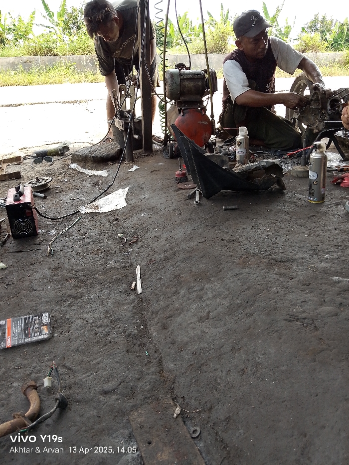 The image shows two men working on a motorcycle in a garage. The men are both wearing casual clothes and are bent over the motorcycle, which is partially disassembled. There is a compressor, tools, and a variety of other objects scattered around the garage. The garage is located in Kecamatan Anjatan, Indonesia, which is located at the coordinates -6.413, 107.953. The floor of the garage is made of concrete and is dirty. There is a piece of plastic sheeting on the ground next to the motorcycle. The men appear to be focused on their work and are interacting with each other. The environment is messy and cluttered. The time is 14:05 on April 13, 2025.