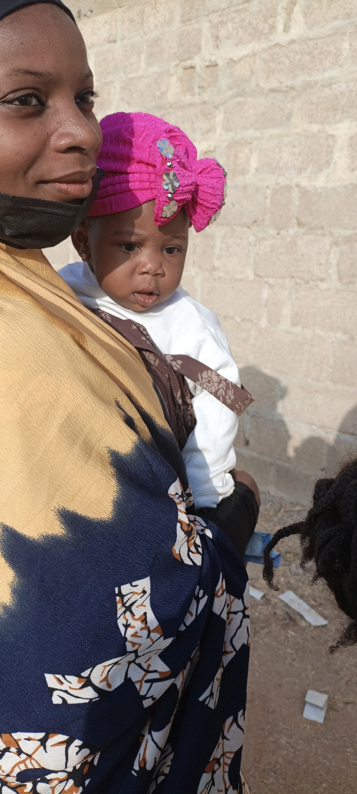 A woman wearing a black mask holds a baby in a carrier. The baby wears a bright pink floral-embellished head covering. The woman wears a yellow and blue patterned garment. The baby has a slight smudge of something near their mouth. Background shows a light brown brick wall and dirt ground with debris. A person with dark dreadlocks stands on the right side of the image.