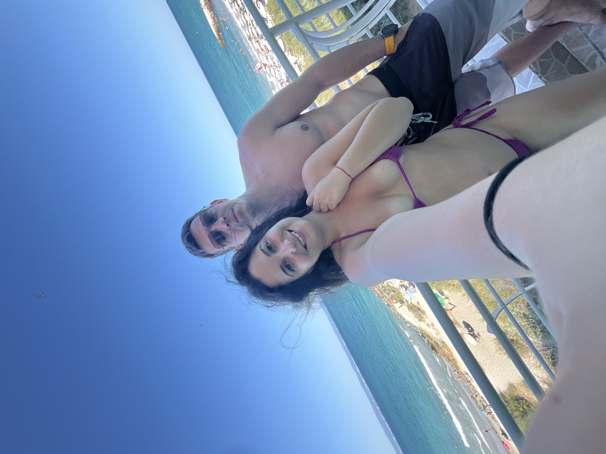 A selfie is taken by a woman in a purple bikini, with a man in swim trunks standing behind her. They are on a balcony overlooking a beach and the Black Sea in Pomorie, Bulgaria. The woman is smiling broadly, her dark hair windswept, while the man looks toward the camera with sunglasses on. The beach below is populated with sunbathers, umbrellas, and lounge chairs. The sky is clear and blue, with a distant kite visible. The railing of the balcony is white and metal.