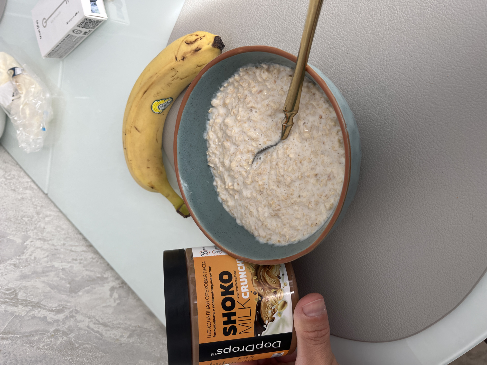 This indoor overhead shot captures a breakfast setting on a light-colored, possibly glass, table with a gray placemat. The primary subjects include a bowl of oatmeal, a banana, and a jar of chocolate peanut butter held by a human hand.
A light blue bowl with a brown rim contains creamy oatmeal, and a golden spoon is partially submerged within it. Lying next to the bowl is a ripe yellow banana, which has a "Village" brand sticker on its skin. In the lower right foreground, a hand holds a jar of "DopDrops™" brand chocolate peanut butter. The jar's label is orange and brown, featuring text in both Russian and English. The Russian text identifies it as "ШОКОЛАДНАЯ ОРЕХОВАЯ ПАСТА" (Chocolate Nut Paste) and highlights "Антиоксиданты и полезные жирные кислоты" (Antioxidants and healthy fatty acids). Below this, the English text reads "SHOKO MILK CRUNCH" and "Chocolate Peanut Butter". An expiry date, "08.02.26", is also printed on the label.
In the upper left portion of the image, there is a white box, possibly a supplement or medication, displaying "Iron (Fe)" and "30 capsules" along with QR codes and other small print. Next to the box, a small, clear plastic wrapper contains an indistinct white object. The bright and even lighting suggests the photograph was taken during the daytime. The use of Russian text on the food product and the supplement box is consistent with the provided location context of Sochi, Russia. The arrangement of items implies preparation for or consumption of a meal, likely breakfast, with the potential addition of the chocolate peanut butter to the oatmeal.