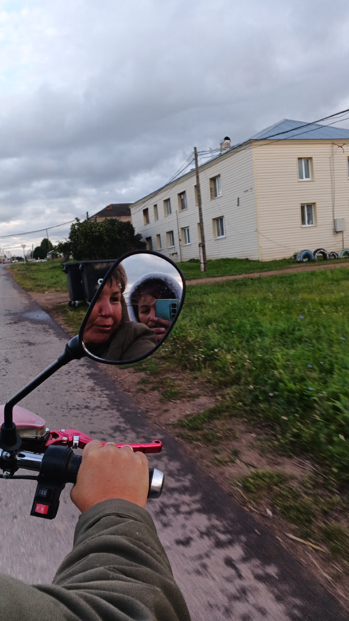 An eye-level, first-person perspective shot captures a hand operating the throttle of a red and black motorcycle, with a portion of the rider's olive-green jacket visible. The motorcycle's side mirror reflects two individuals: one with short, brown hair and another with dark hair and a grey hooded jacket, holding a turquoise smartphone to take a selfie. They appear to be riding on a paved road in the village of Podgornoye, Russia, judging by the style of the beige apartment building with a blue roof in the background, which is typical of rural Russian architecture. Large black trash bins are situated on the left side of the road, and lush green grass lines the path. The sky is overcast with heavy grey clouds, indicating a potentially gloomy or stormy day.