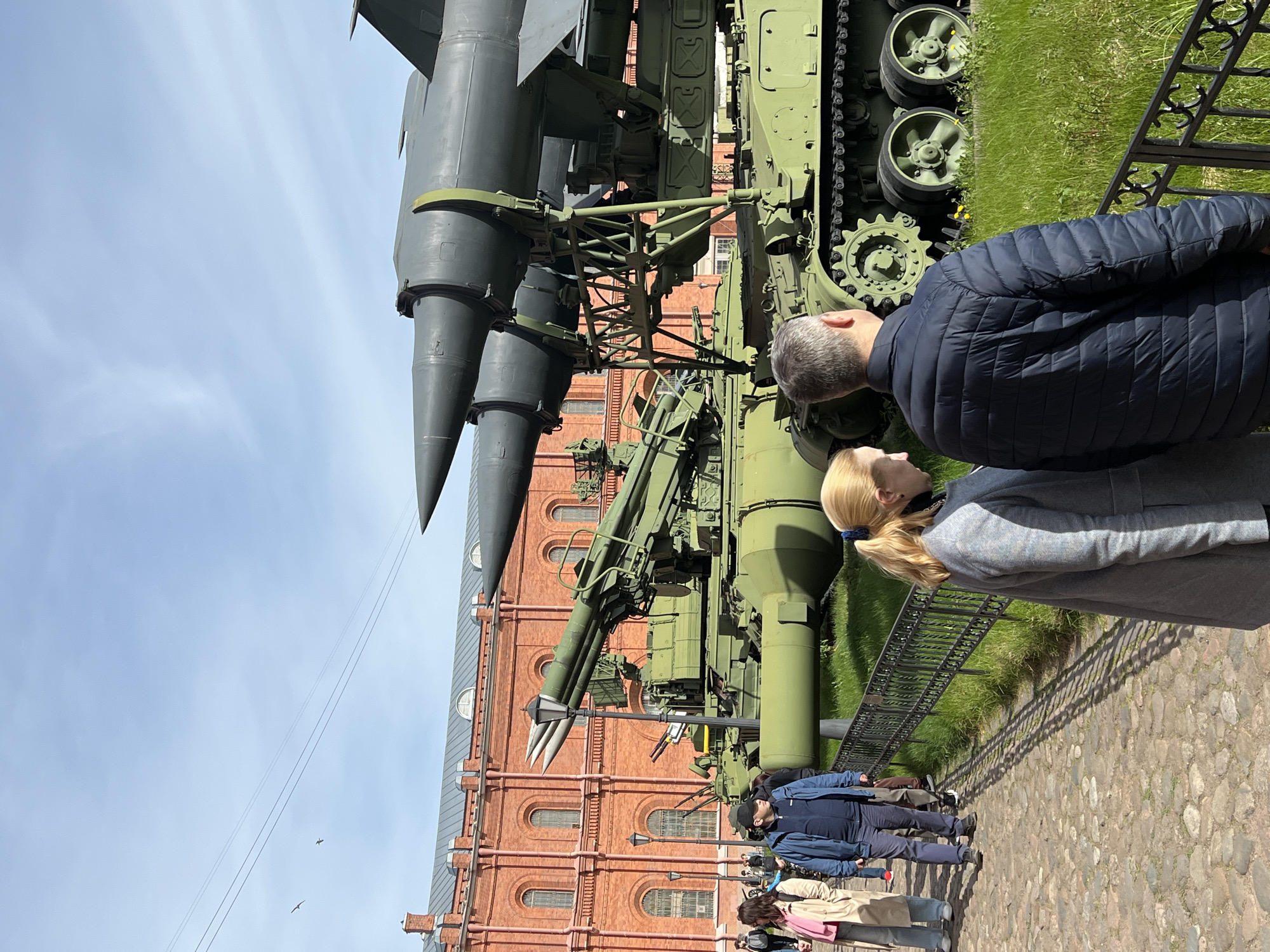 The image shows a mobile surface-to-air missile system, likely an S-75 Dvina, exhibited outdoors near a brick building. People are looking at the missile system. The location appears to be in Saint Petersburg, Russia, possibly near the Artillery Museum. The missile is painted dark green/gray, mounted on a tracked vehicle. Several individuals stand on a paved path, observing the equipment. The sky is pale blue with some overhead wires visible.