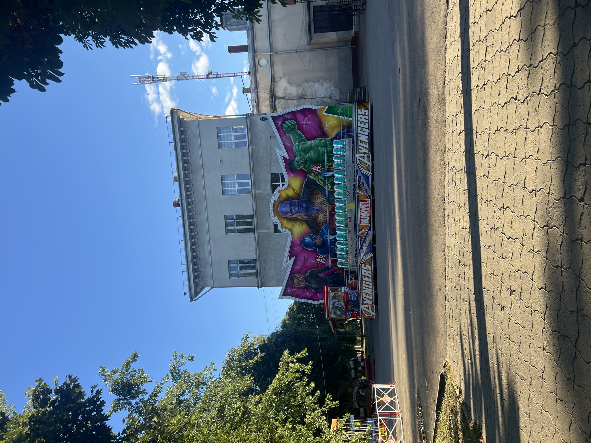 A bright, sunny day in the city of Sheptytskyi, Ukraine, showcases an outdoor amusement park attraction featuring Marvel's Avengers characters. The central focus is a ride with a vibrant backdrop depicting Thanos, Hulk, and Captain America. Black Widow is also visible on the left side of the ride. The ride itself has multiple rows of seats, appearing to be designed for children or a family-friendly experience. In the background, a light-colored, multi-story building stands prominently, with a cell tower visible on its right. To the left of the building, lush green trees provide a natural contrast to the urban setting. The ground is paved with interlocking stones, casting long shadows from the trees. To the far left, a smaller amusement ride, possibly a carousel or kiddie ride, is partially visible. The scene suggests a lively and recreational atmosphere in an urban park.