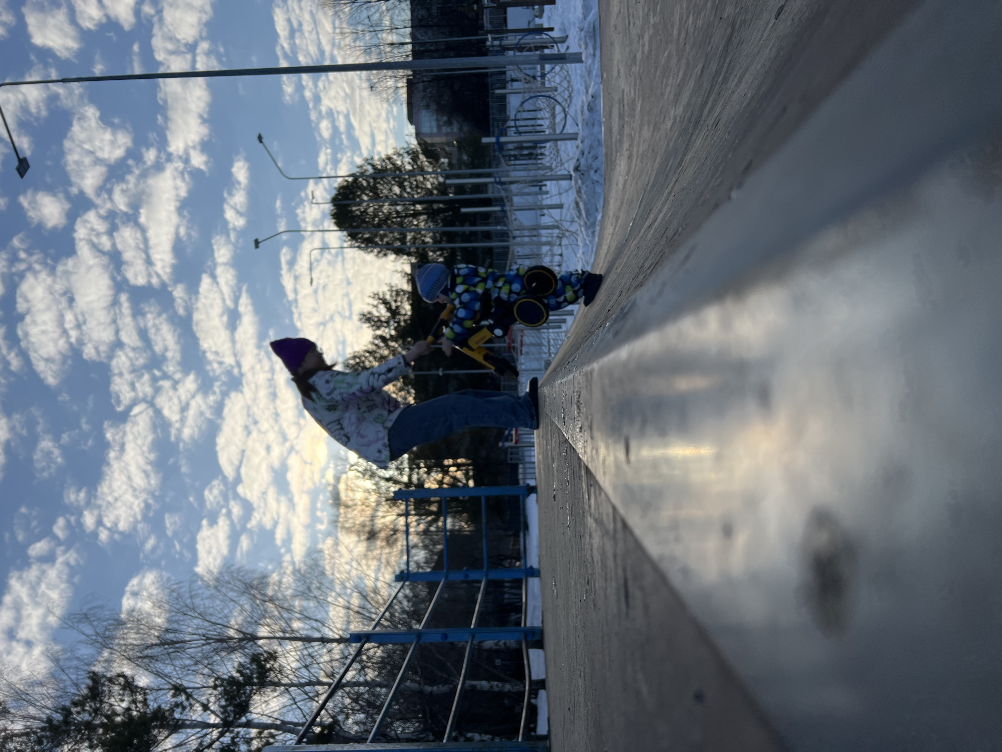 The photograph depicts a scene in what appears to be a recreational area in Chaikovsky, Russia. Two individuals are present: an adult wearing a purple hat and light-colored patterned outerwear, and a child in a patterned snowsuit and blue hat. The child is seated on a yellow snow scooter. The adult seems to be assisting the child, possibly preparing for a ride down a snow-covered slope or ramp. The location includes a metal railing alongside the slope, with bare trees and a cloudy sky visible in the background. Streetlights are also visible. Other elements of the park can be seen further back, partially obscured by snow.