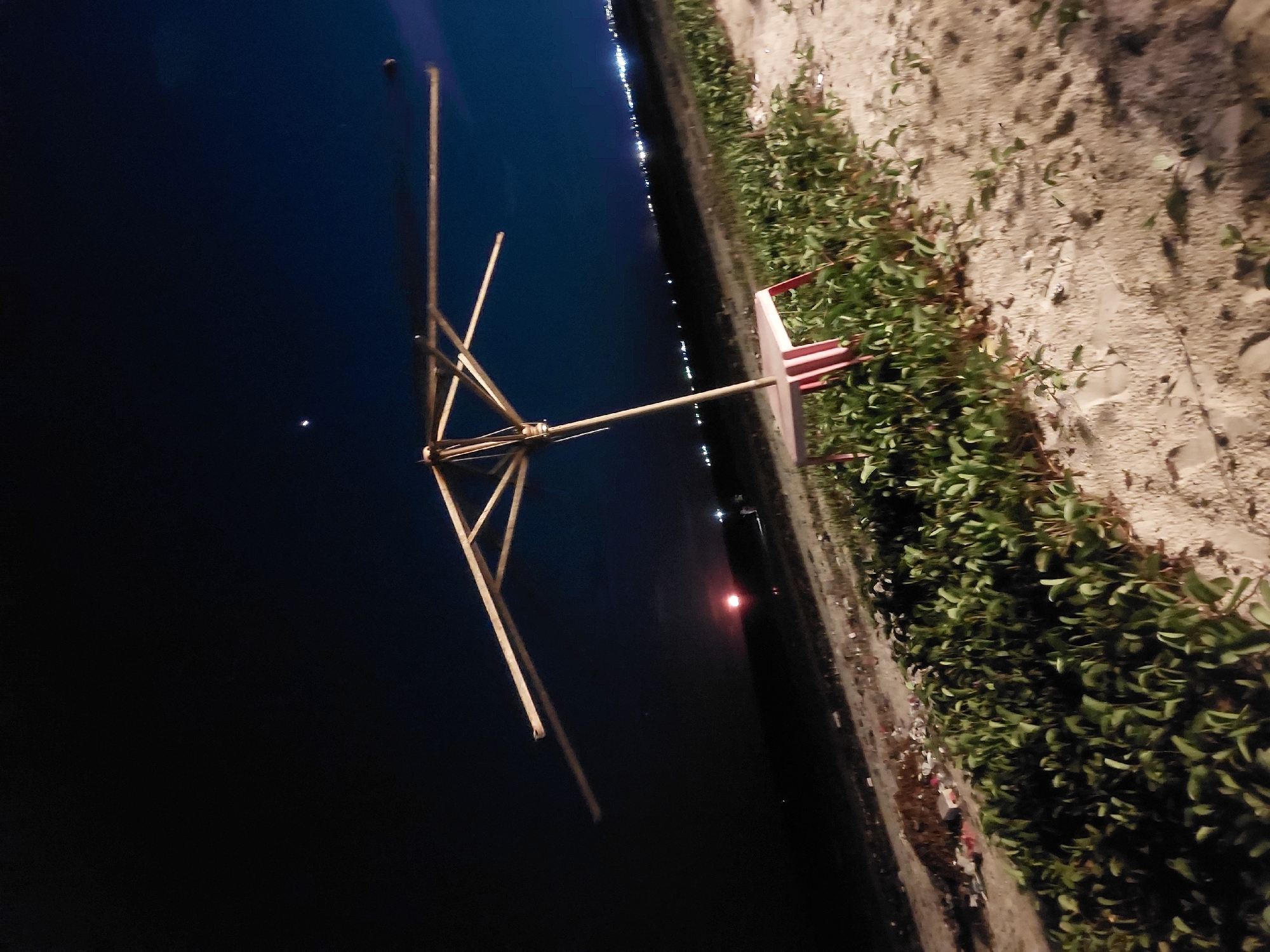 At night, the scene shows a body of dark water, reflecting lights from the distant city of Dar es-Salaam, Tanzania. On the edge of the water is a vegetation-covered bank, transitioning to a sandy or concrete surface. A structure resembling a dilapidated wind vane or similar device stands tall. It's made of wooden or bamboo poles lashed together, with a red and white fin at its base.