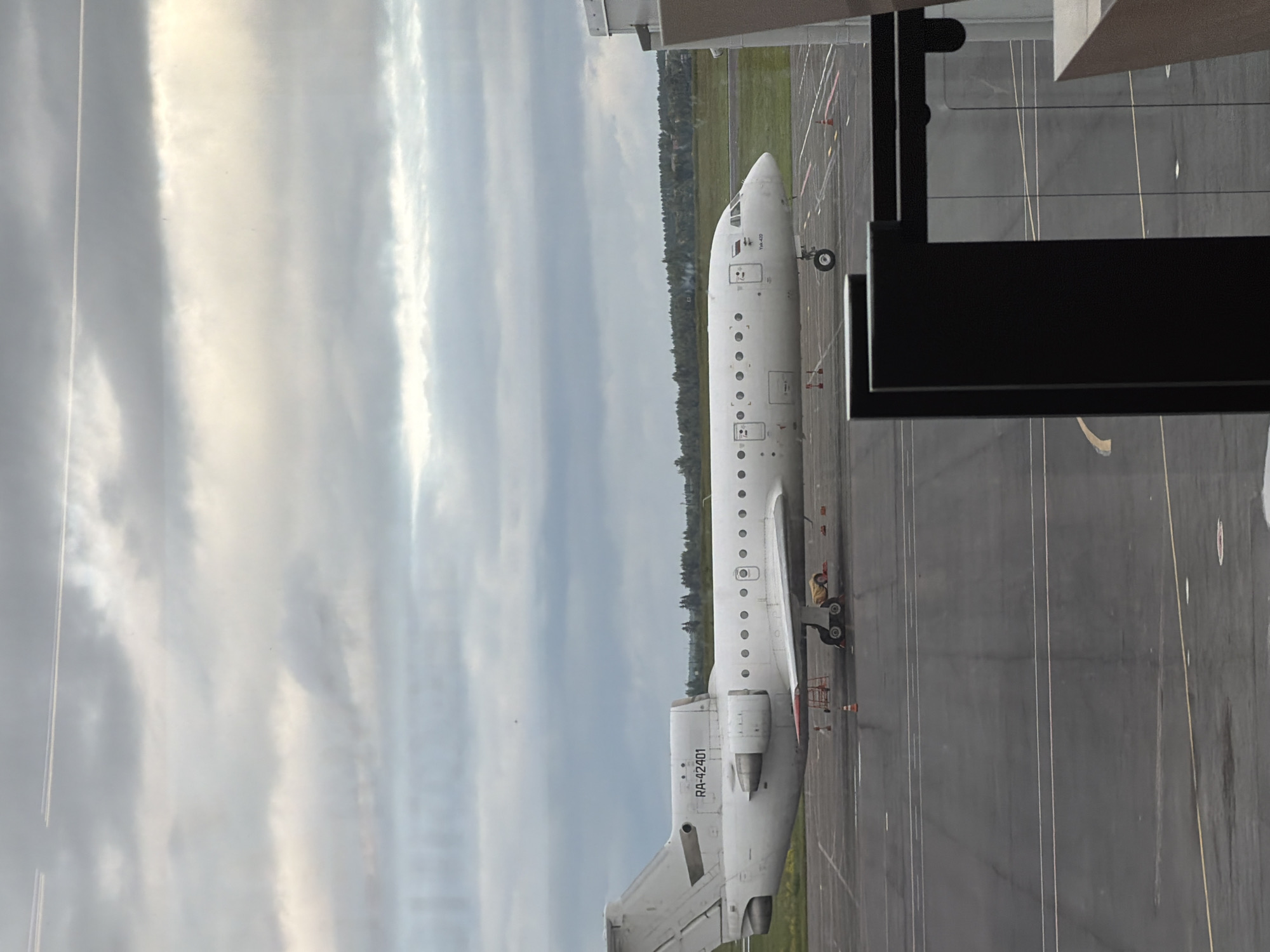 The image depicts a static white Yakovlev Yak-40 trijet aircraft, registration RA-42401, parked on a dark asphalt apron. The aircraft is oriented horizontally, facing right, with its landing gear deployed. Three engines are visible on the tail section, a characteristic of the Yak-40. Small circular windows line the fuselage. Orange traffic cones and support equipment are positioned near the aircraft's undercarriage and wings. The apron features white and yellow painted markings, indicating pathways or parking positions.
Beyond the apron, a strip of green grass separates the tarmac from a dense line of green trees. The distant background shows a flatter, lighter green landscape under a heavily overcast sky. The sky is filled with layered grey and white clouds, with two distinct white contrails from high-flying aircraft visible in the upper portion. The image appears to be taken through a window, as evidenced by partial reflections and dark structural elements of a building frame visible in the lower left and right foreground. The scene is located in Новое Мартьяново, Russia. No people are visible.