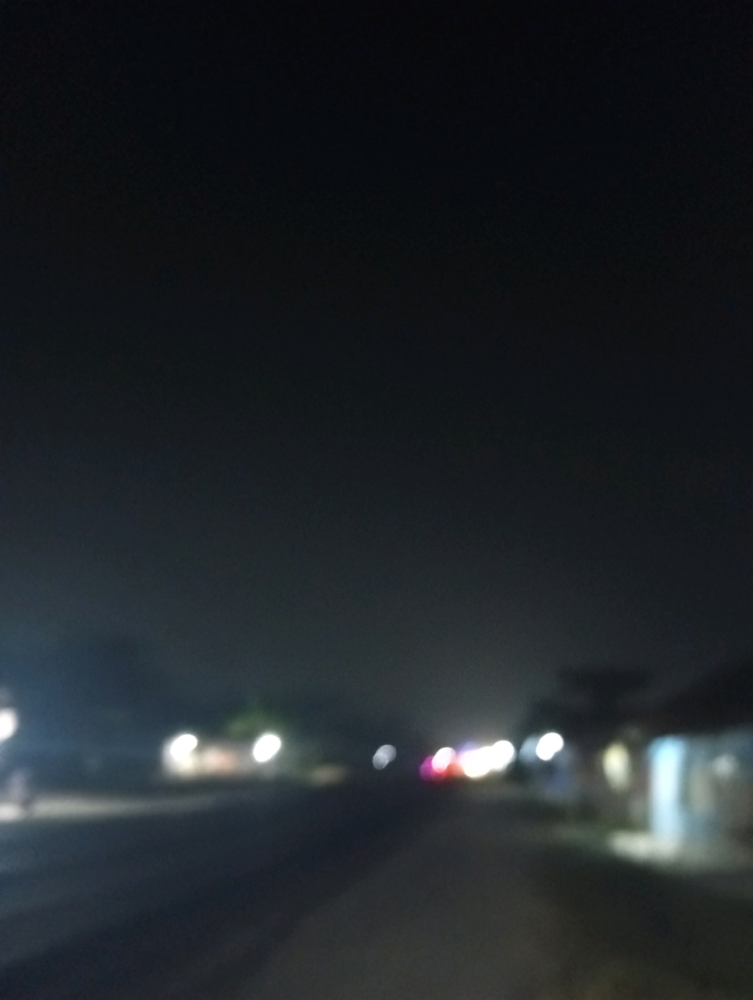 The image is a blurred, night-time photograph taken outdoors in Kubang Jaya, Indonesia. It appears to show a road with some indistinct structures and lights on either side, suggesting an urban or semi-urban environment. The darkness of the sky indicates it is likely late at night. There are no discernible people, subjects, or specific objects clearly visible due to the blurriness. The overall scene is dimly lit with scattered bokeh effects from lights.