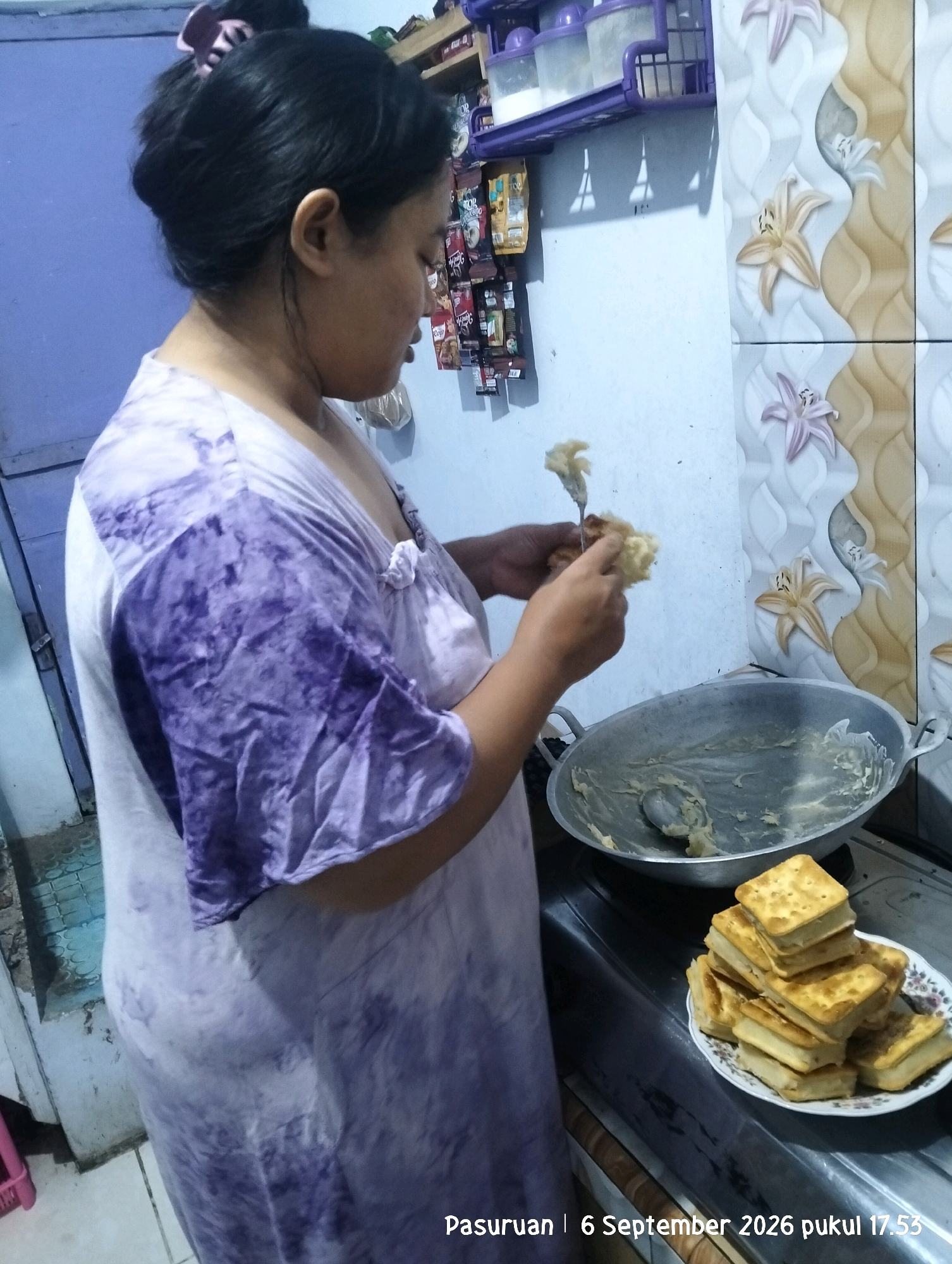 The image depicts a woman in a tie-dye purple and white dress standing in a kitchen. She is holding a fork with a piece of food on it, seemingly preparing to eat or arrange it. In front of her, on a stovetop, is a large metal pan with remnants of food and a spoon, and to the right, a plate of stacked square crackers. The background shows kitchen cabinetry with items on shelves, and a tiled wall with floral patterns. The scene appears to be indoors, likely in a home kitchen. A timestamp at the bottom of the image indicates "Pasuruan | 6 September 2026 pukul 17.53," suggesting the location is Pasuruan, Indonesia. Warung Dowo is a village within the Pasuruan Regency, so this image could be from that area.