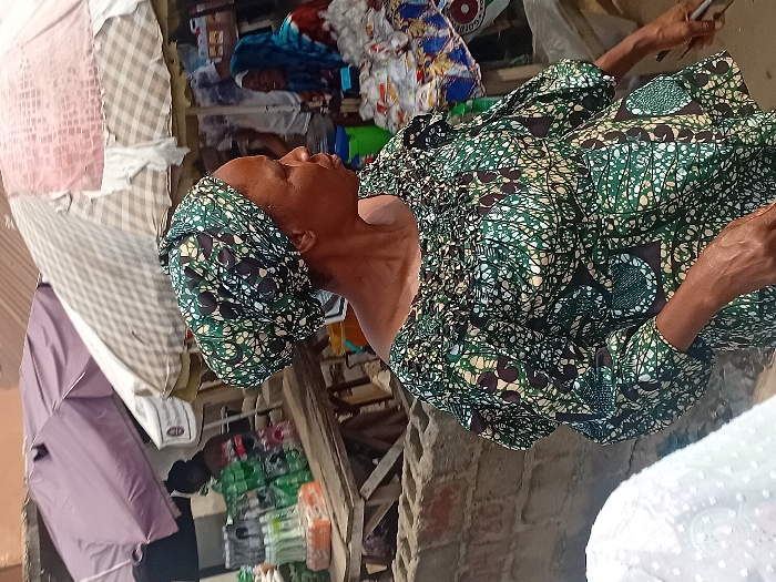 An older woman wearing a green and black patterned dress and headwrap is seen from the chest up, holding what appears to be a cellphone in her right hand. The setting looks to be inside a cluttered building, with fabrics piled in the background. A light pink umbrella and stacked packages are visible in the lower-left corner. The image has been rotated counter-clockwise, causing a tilted orientation.