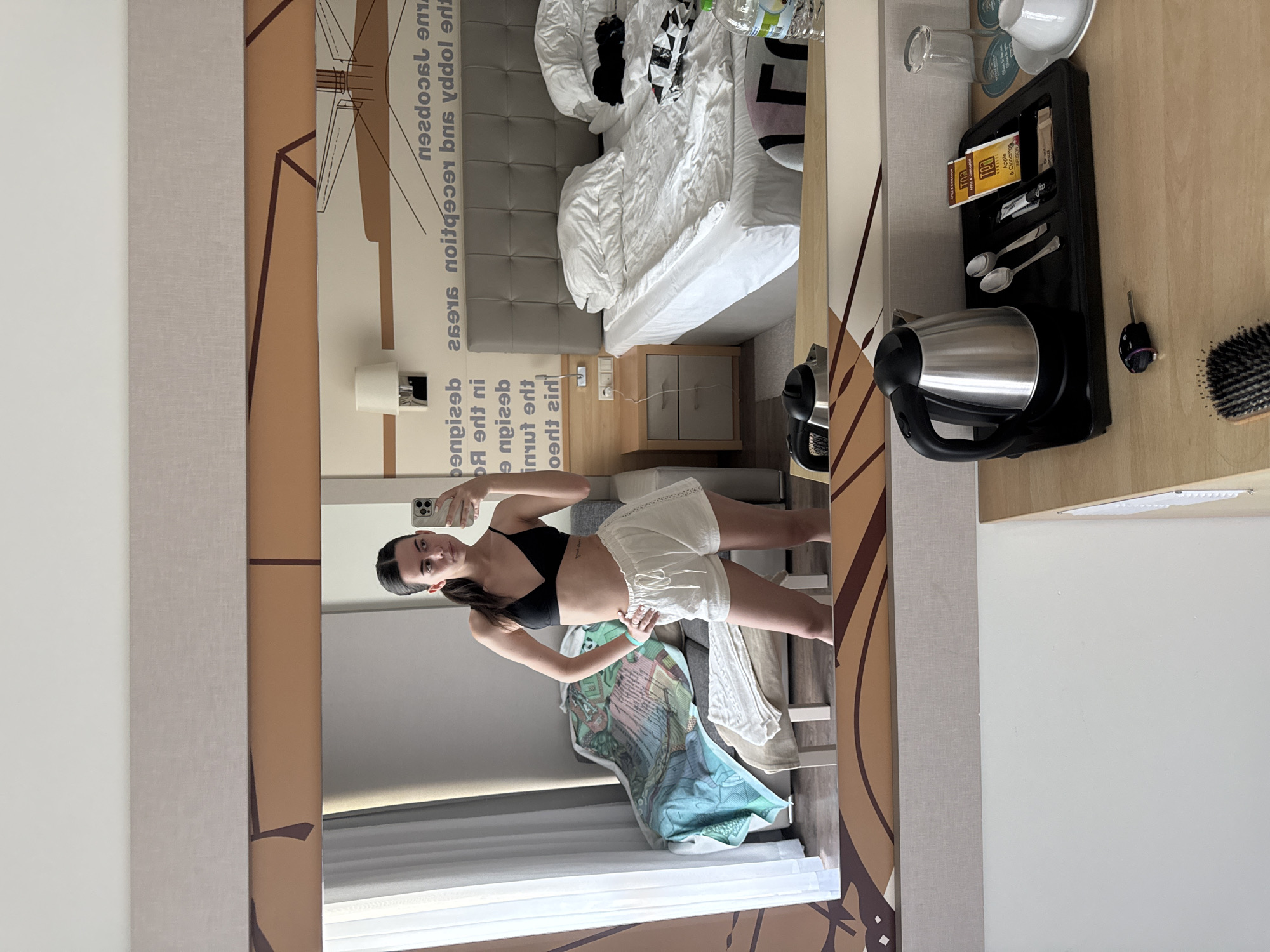 A young woman is taking a mirror selfie in a hotel room. She is wearing a black bikini top and white shorts. The room has a large bed, a nightstand, and a chair with a blanket on it. There is also a desk with a kettle, tea bags, spoons, and a glass. The woman is standing in front of a mirror, and her reflection is visible in the image. The room appears to be clean and well-maintained. The overall impression is one of a comfortable and relaxing hotel stay.