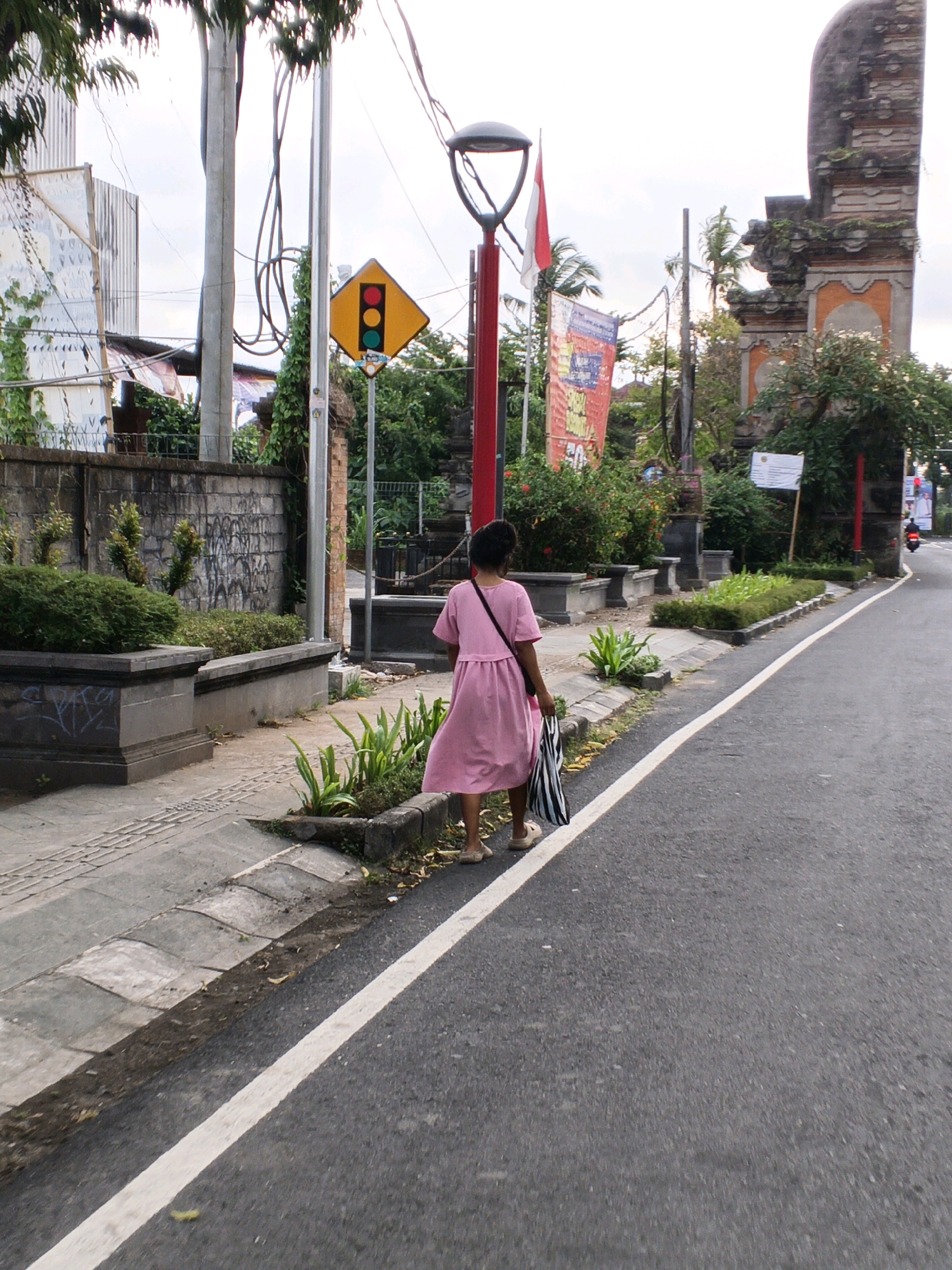 A person wearing a pink dress walks away from the camera on a paved sidewalk next to a road in Blahbatuh, Indonesia. The person is carrying a black and white striped bag over their shoulder. On the left side of the image, there is a stone wall covered in graffiti, and beyond that, a white building with some decorative elements. A traffic light with red, yellow, and green lights is visible on a pole, along with several wires and flags. On the right side of the image, there is an ornate stone archway with weathered brickwork, surrounded by greenery. The road has a white line painted down the center.