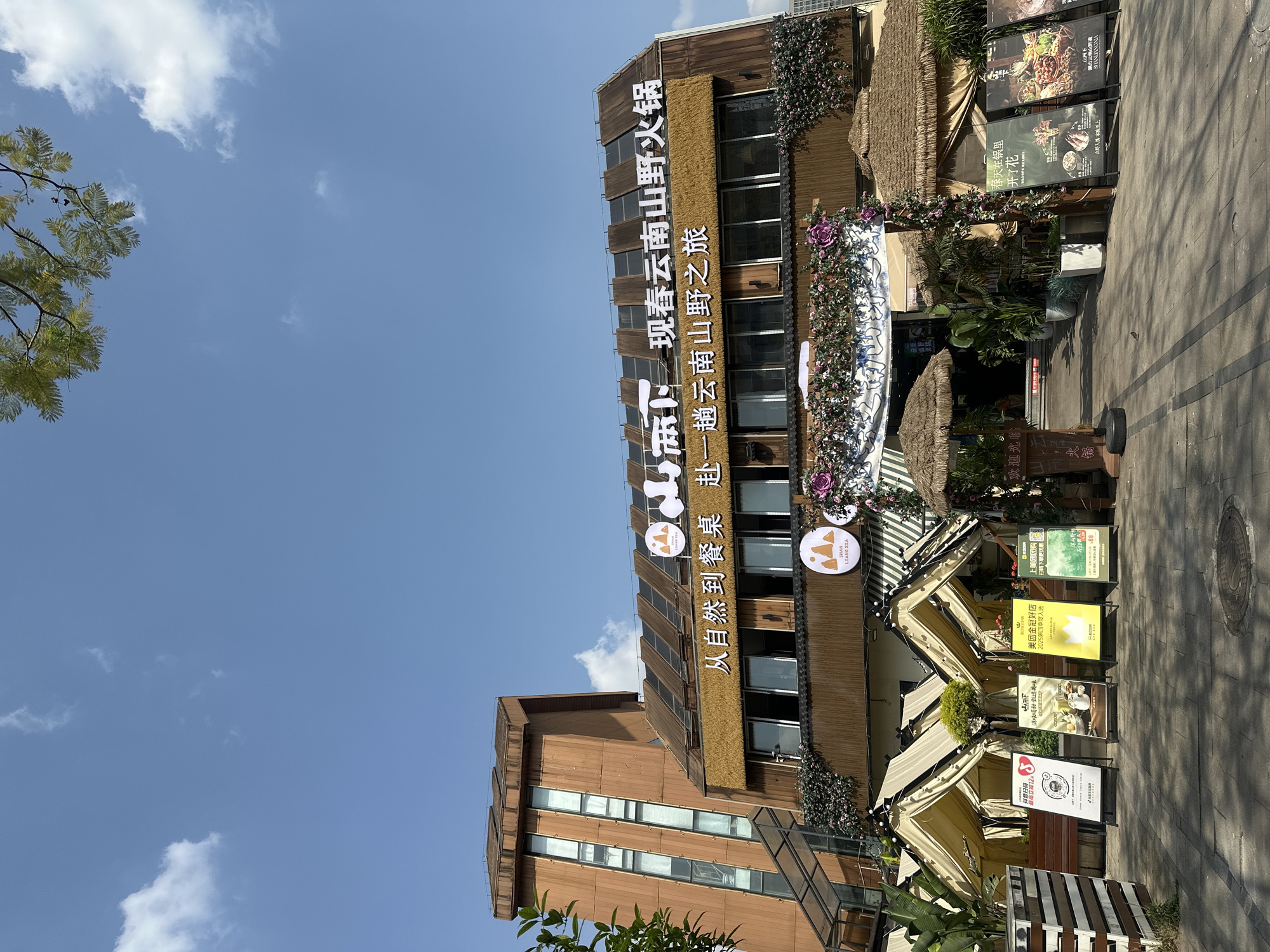 This is a bright daylight image capturing the exterior of a restaurant building in Chongqing High-tech Zone, China, under a clear blue sky with scattered white clouds. The building features a distinctive, rustic aesthetic with wooden facades, horizontal window panels, and straw or thatch-like accents above the windows, giving it a natural, "wild" or traditional feel. Floral decorations, primarily purple and green foliage, adorn the building and an archway leading into the establishment.
The main building sign, prominent on the facade, reads "山下现春云南山野火锅" (Mountain Foot Spring Yunnan Wild Hotpot) and a slogan "从自然到餐桌赴一趟云南山野之旅" (From nature to the dining table, embark on a Yunnan wild journey). A circular logo with text "SHAN LIANG XIA" is also visible. Below, on the ground level, several awnings extend outwards, providing shade over what appears to be an outdoor seating area or entrance.
A series of standing signs on the pavement advertise the restaurant's offerings and promotions. Text on these signs includes "欢迎光临" (Welcome), "抖音扫码 最高立减12" (Douyin (TikTok) scan code, save up to 12), "海味吸甜·甄选美味" (Sweet seafood · Selected delicacies), "美团金冠好店 2025第四季度入选" (Meituan Gold Crown Excellent Store, selected for Q4 2025), "上美团团购 扫码下单更优惠" (Go to Meituan Group Buy, scan code to order for more discount), and a slogan about hotpot: "春天在锅里 开了花" (Spring blossoms in the hotpot). Another sign mentions "山下 湖出云南山野魂" (Mountain Foot, the lake brings out the soul of Yunnan wilderness). Various plants in pots are placed around the entrance, enhancing the natural theme. The pavement is made of concrete tiles, and shadows indicate a bright, sunny day.