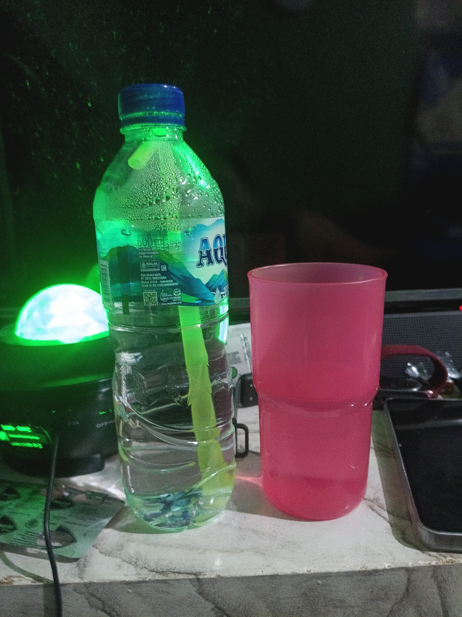 The image depicts a still life composition on a textured, light-colored surface, possibly a countertop or table. In the foreground, a transparent plastic bottle of "Aqua" mineral water, illuminated by a green light source from the left, stands prominently. The bottle is covered in condensation, indicating it is cold. Inside the bottle, a green straw is visible, submerged in the water. To the right of the bottle, a bright pink, opaque plastic cup filled with a clear liquid is positioned.

In the background, a dark, indistinct area suggests a room or a cluttered surface. A glowing, faceted object emitting a bright white and green light is visible to the left of the bottle, contributing to the overall illumination and setting a somewhat moody atmosphere. The presence of what appears to be electronic equipment, such as a keyboard or speaker grille with green indicator lights, is hinted at behind the bottle and cup. A black cable extends from the left side of the frame towards the bottle. The overall scene is dimly lit, with the green and pink colors creating a strong contrast. There are no people or animals visible in the image, and no specific actions or interactions are depicted. The context of "Sukamekar, Indonesia" is not visually discernible from the image itself.