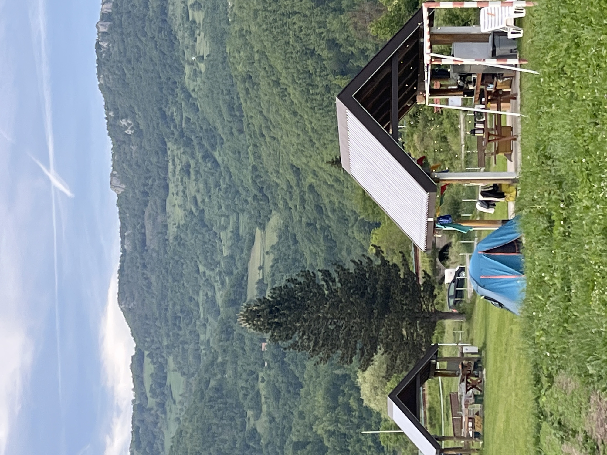 The scene shows a rural landscape in Polja, Montenegro. A lush, green mountain dominates the background under a blue sky with faint contrails. In the foreground, a grassy area is populated with structures. There are two angular roofed pavilions, both with metal beams and posts, and light-colored slatted roofs. One pavilion is larger and contains wooden furniture, while the smaller one is partly obscured. A blue tent is visible in the midground. A large tree stands between the structures, with dense foliage. Tall green vegetation lines the right edge of the frame. There are no people immediately visible.