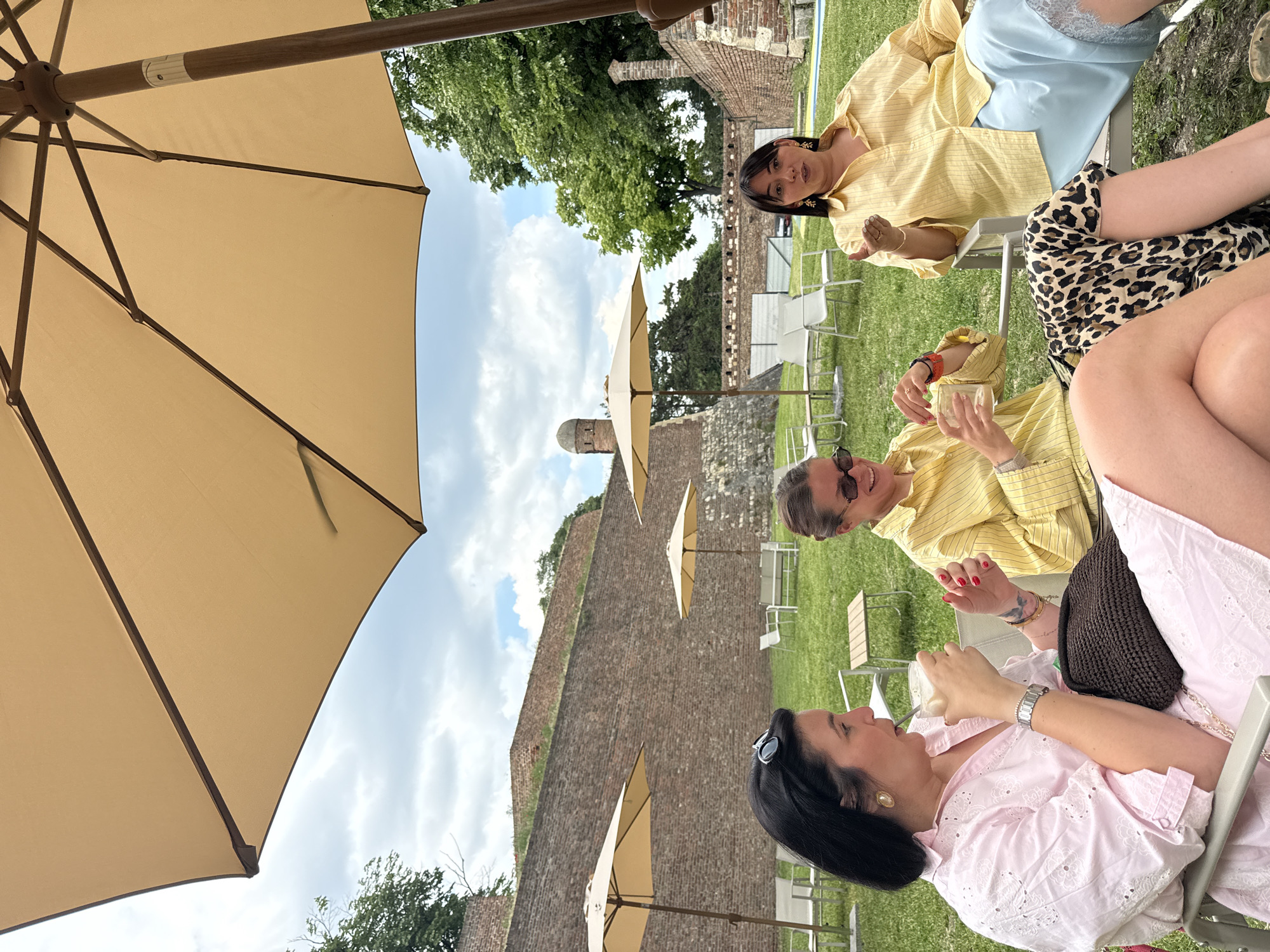Three women sit outdoors on chairs in Belgrade, Serbia. The women are dressed casually in light-colored clothing. A fourth person is partially visible. There are umbrellas providing shade. A brick wall is in the background. A tower is visible in the distance. The women appear to be interacting and drinking beverages.