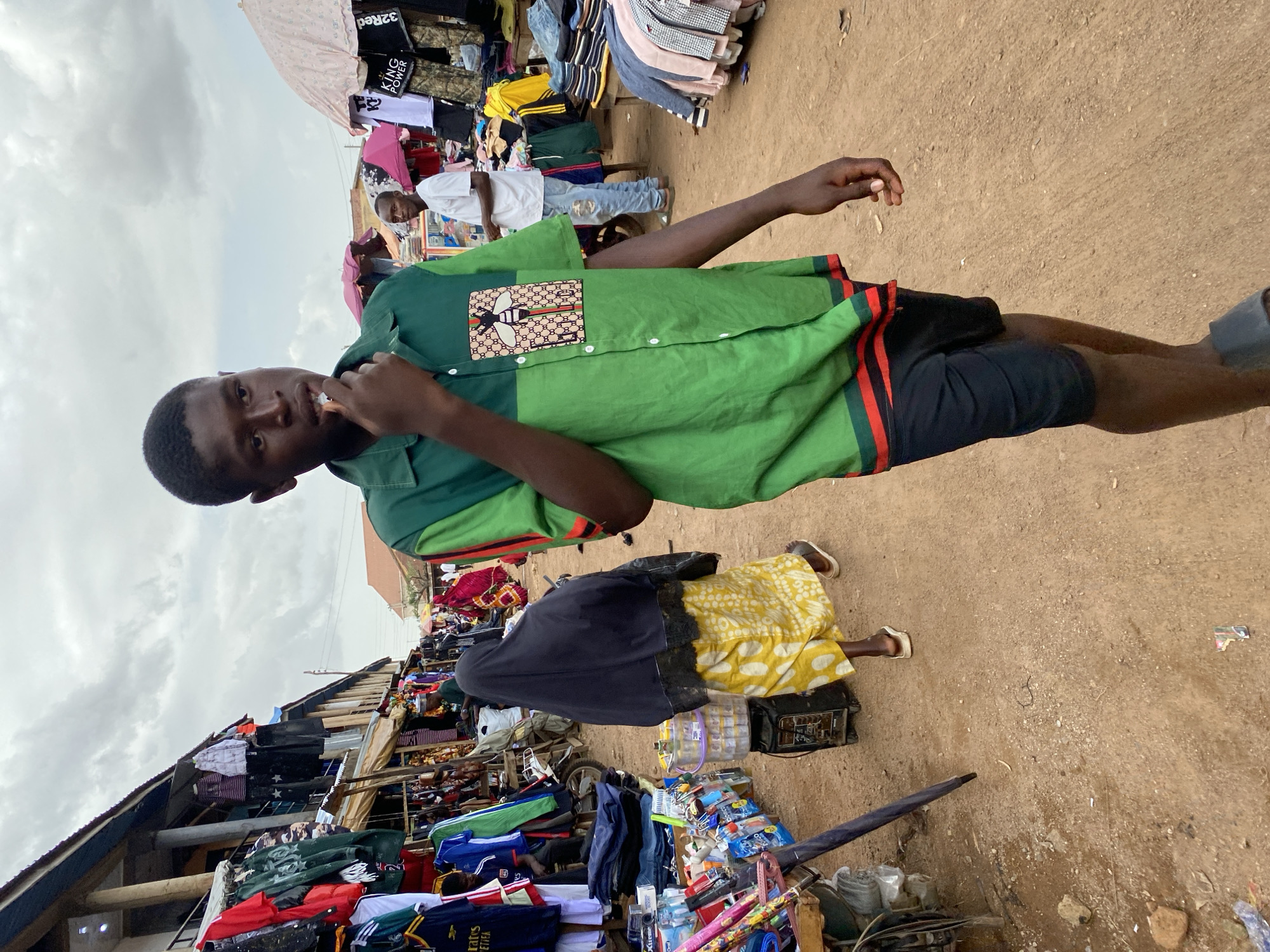 In Bukuru, Nigeria, a bustling outdoor market is depicted. A young Black man, wearing a green short-sleeved shirt with a bee emblem and black shorts, walks towards the viewer, touching his mouth with his fingers. Behind him, a woman in a black head covering and a yellow patterned skirt walks away, and other market-goers and stalls are visible. The ground is dirt, and the sky is overcast.
