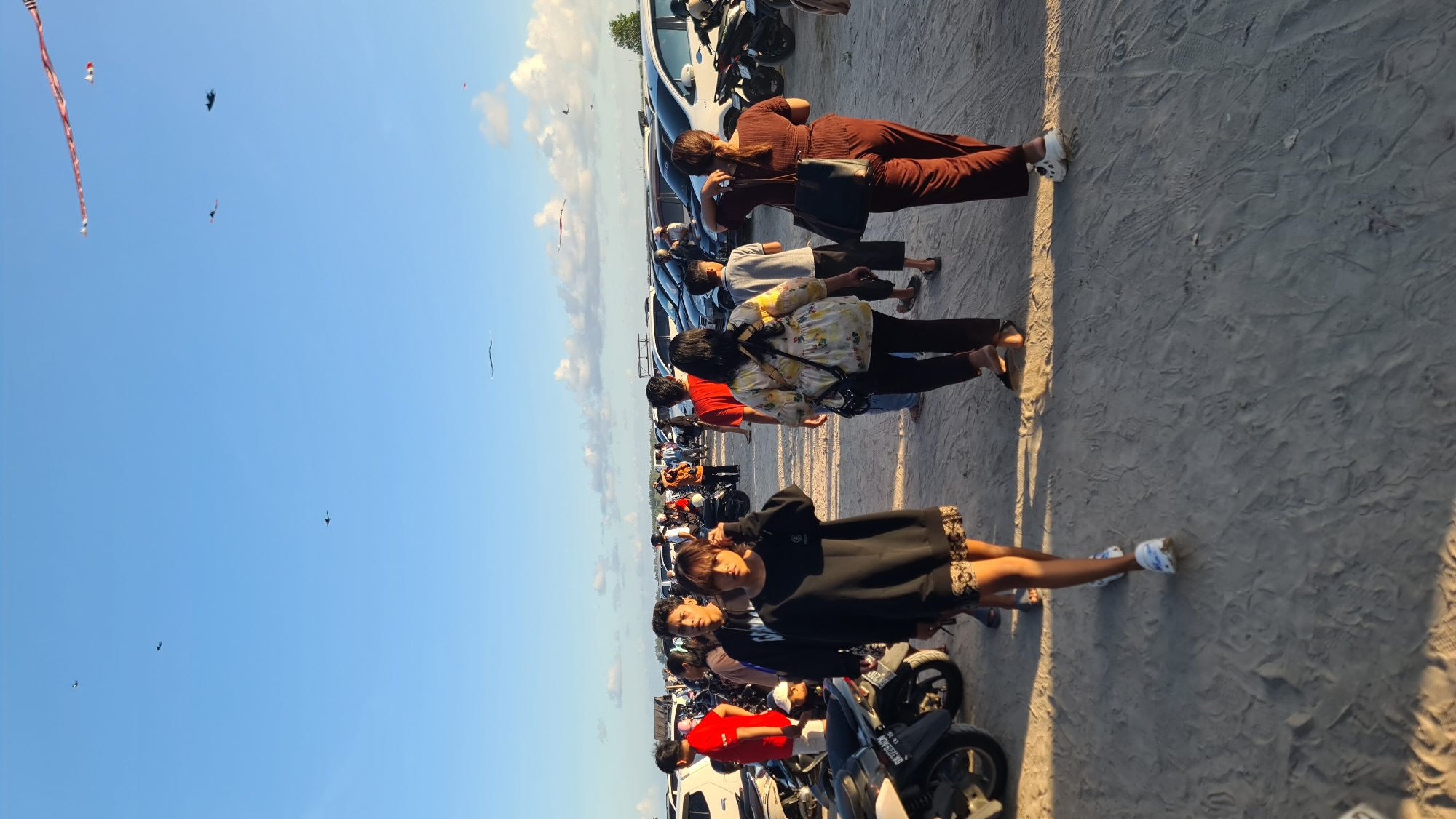 The image depicts a crowd of people gathered on a sandy area, likely a beach or parking lot, with several motorcycles and cars parked in the background. The sky is blue with some scattered clouds. Several kites are visible flying in the sky. In the foreground, a young woman in a black sweatshirt and patterned skirt is walking towards the camera. Behind her, other individuals are walking in various directions. The scene appears to be outdoors in Denpasar, Indonesia, given the presence of parked vehicles and the general atmosphere. The lighting suggests it is late afternoon or early evening.
