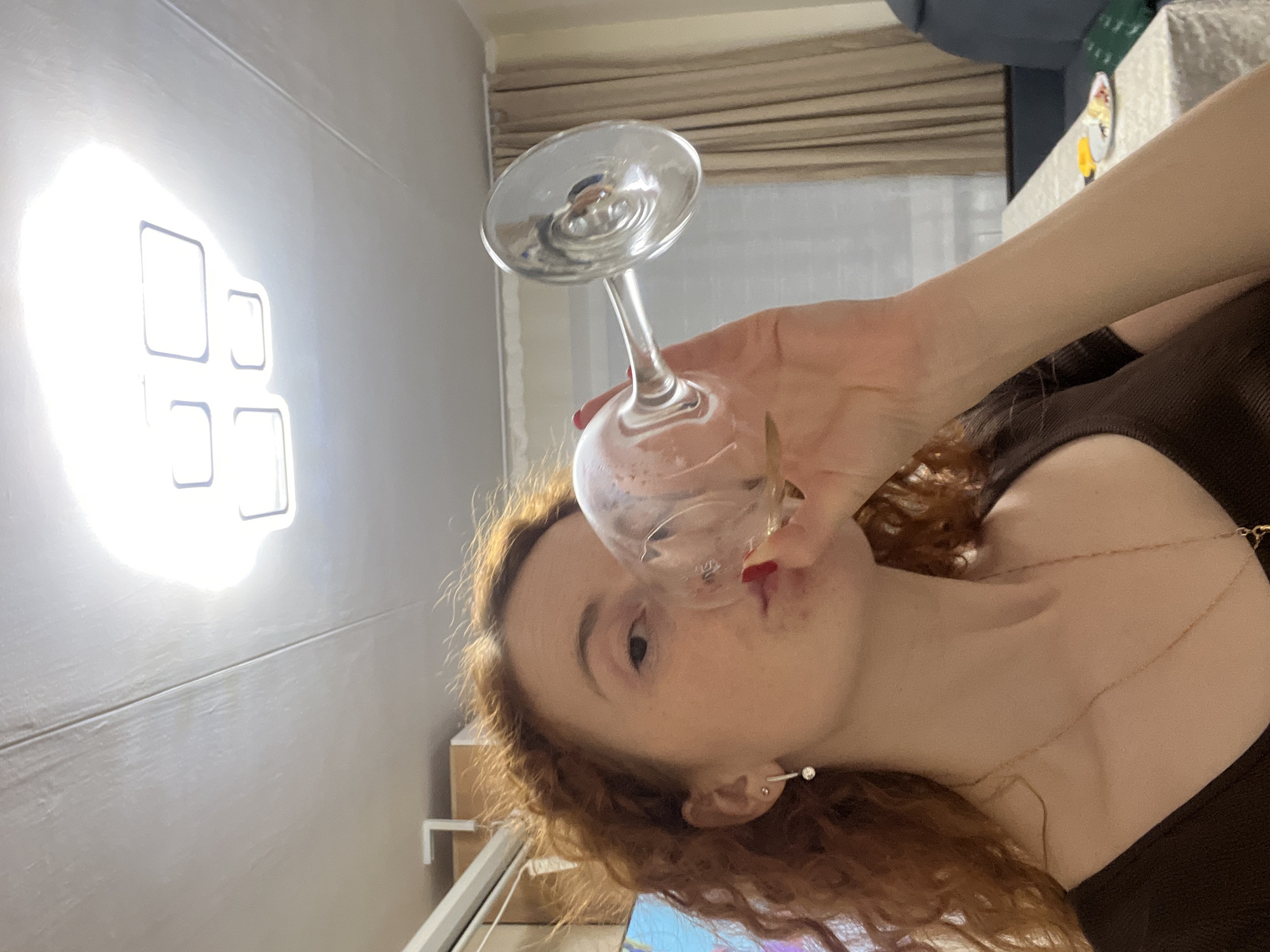 A young woman with red hair is holding a wine glass to her lips, seemingly about to drink. She is wearing a brown top and a delicate gold necklace and earrings. The background includes a beige curtain, a wall with a square pattern, and a glimpse of what might be a table or desk. Based on the details visible, the exact location in the city of Chaikovsky, Russia, cannot be determined from this image alone.
