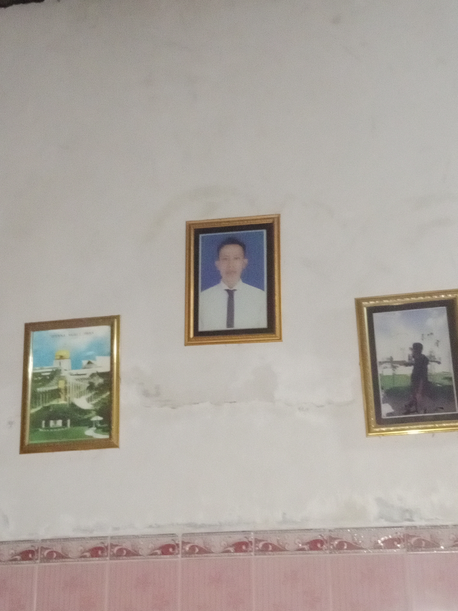 Three framed pictures hang on a white wall. The picture on the left depicts an architectural landscape with a golden dome. The middle picture is a portrait of a man in a white shirt and tie. The picture on the right is a silhouette of a person with arms outstretched. The bottom of the image shows a tiled wall with a decorative border. The scene appears to be indoors.