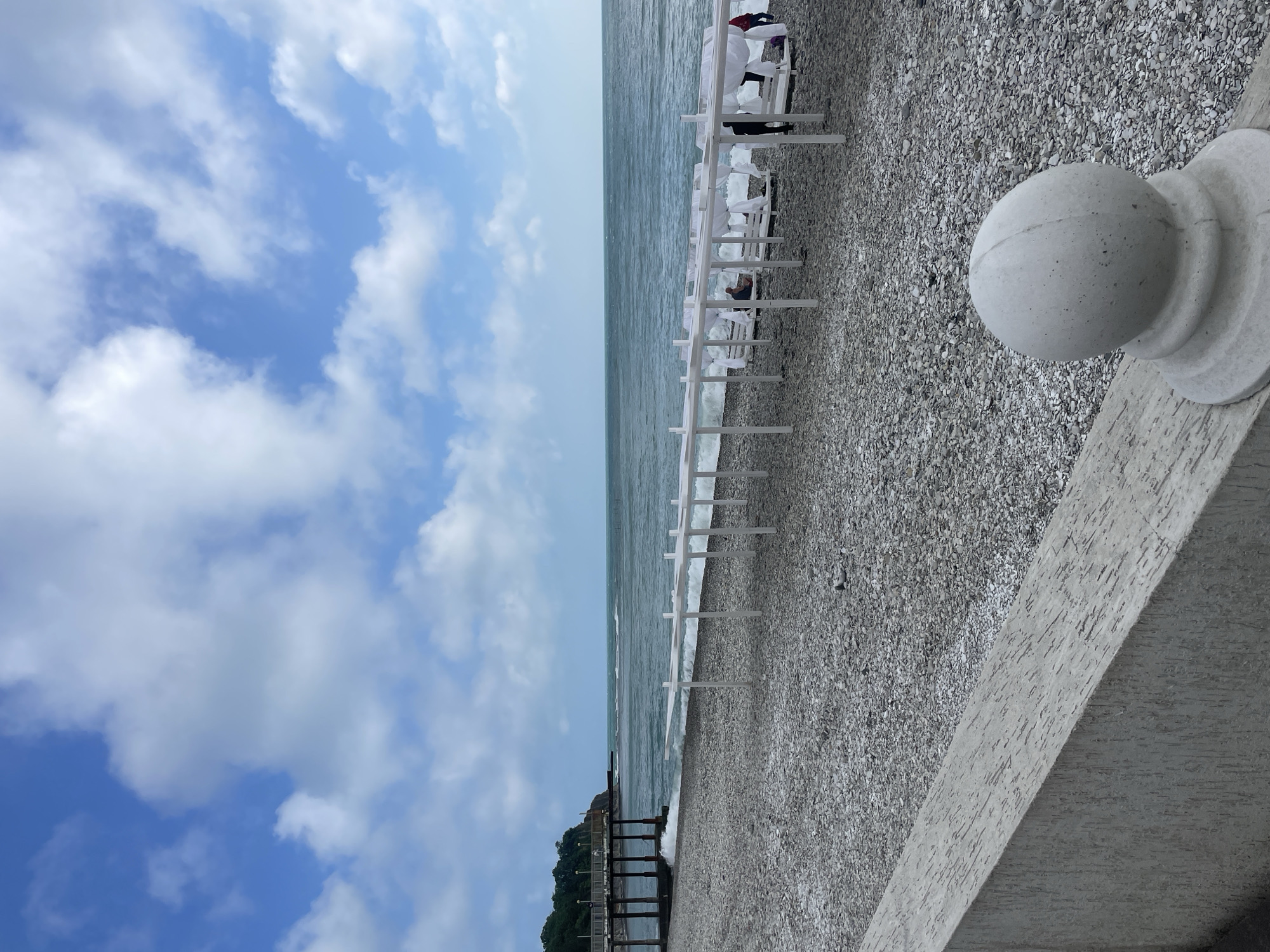 The image shows a pebble beach on a cloudy day in Olginka, Russia. White beach canopies and lounge chairs line the shore. A concrete wall with a decorative sphere is in the foreground. A wooden pier extends into the water. The sky is blue with white clouds. The sea is a dark grey-blue.