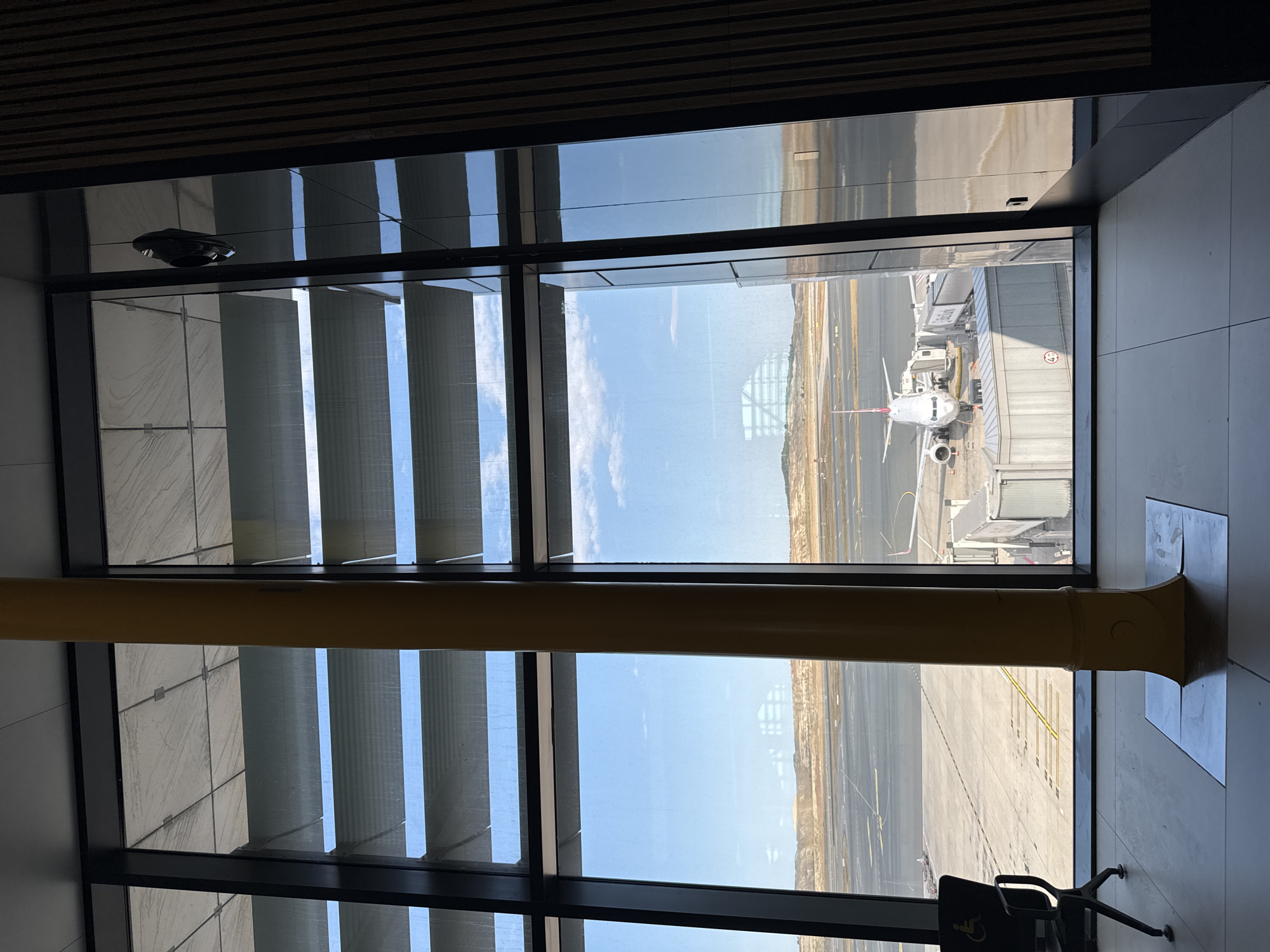 The image shows an interior view of an airport terminal at Istanbul Airport (IST) in Arnavutköy, Turkey. A large yellow pillar dominates the foreground, situated between two large window panels. Through the windows, an airplane is visible parked at a gate, connected by a jet bridge. The tarmac and surrounding airport infrastructure, including a distant runway and arid landscape, are also visible. The sky is clear with scattered clouds. A disabled seating area is visible to the left, and a reflection of a person's shoe hanging from a string is seen in the upper right portion of the window. The interior appears modern with tiled flooring and wooden paneling on the right wall.