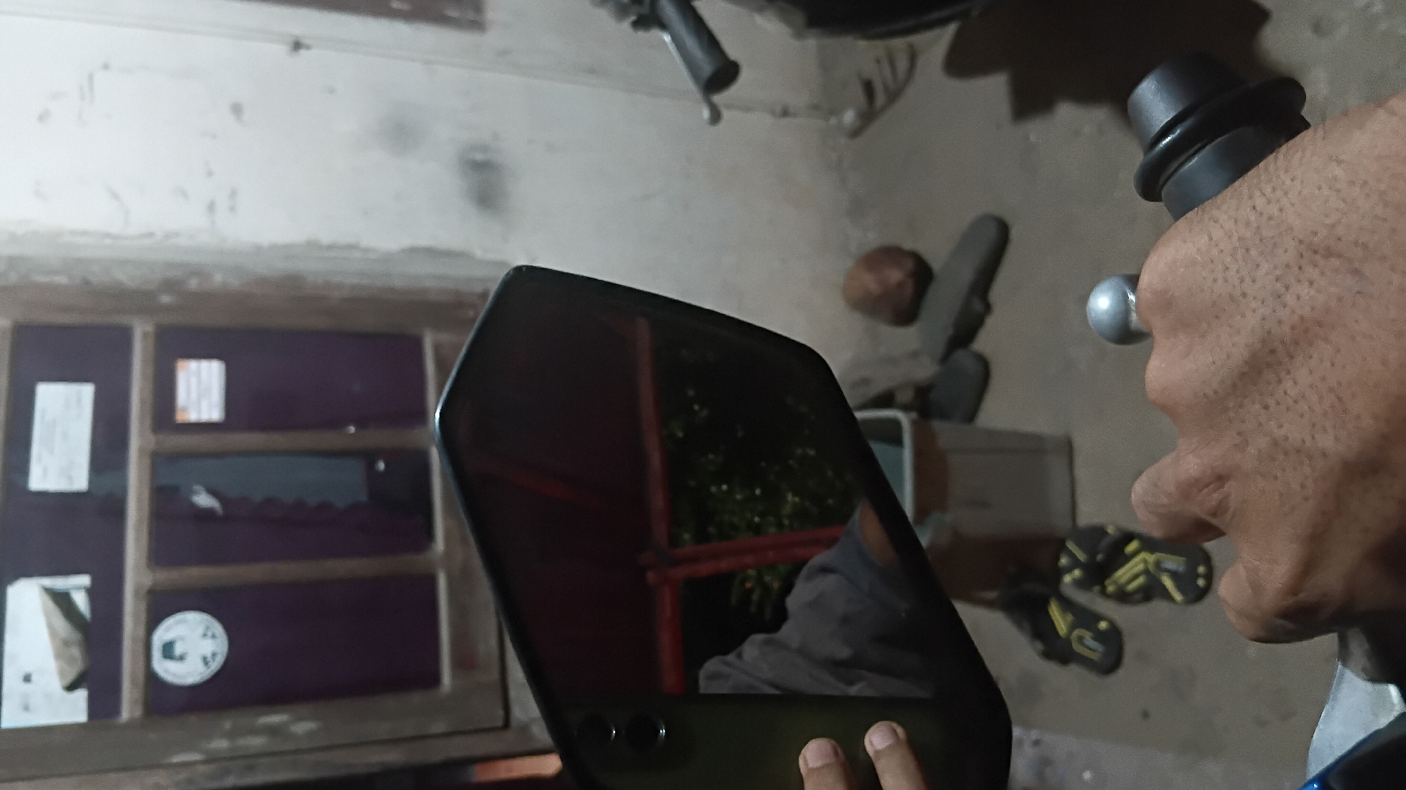 The image captures a first-person perspective of someone holding a motorcycle handlebar, with a side mirror prominently reflecting a portion of their arm and upper body. The reflection also shows a dark-colored object, possibly a door or window frame, with what appear to be several papers or labels affixed to it. The background is dimly lit and suggests an outdoor or semi-outdoor environment, possibly a yard or a pathway adjacent to a building. There are several objects scattered on the ground, including a pair of flip-flops, a smooth, rounded stone, and a rectangular stone or tool. The wall in the background appears to be rough and light-colored, with some smudges or stains. The scene is likely in Prabumulih, Indonesia, given the context provided.