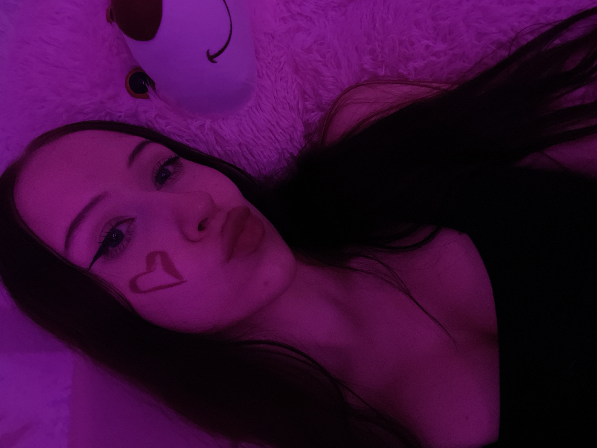 A young woman with long, dark hair is shown in a close-up selfie, illuminated by purple lighting. She has eyeliner and a heart shape drawn on her cheek. She is wearing a black top and is lying down next to a plush toy. The background appears to be a soft, textured surface. There is no information to suggest this image is located in Votkinsk, Russia.
