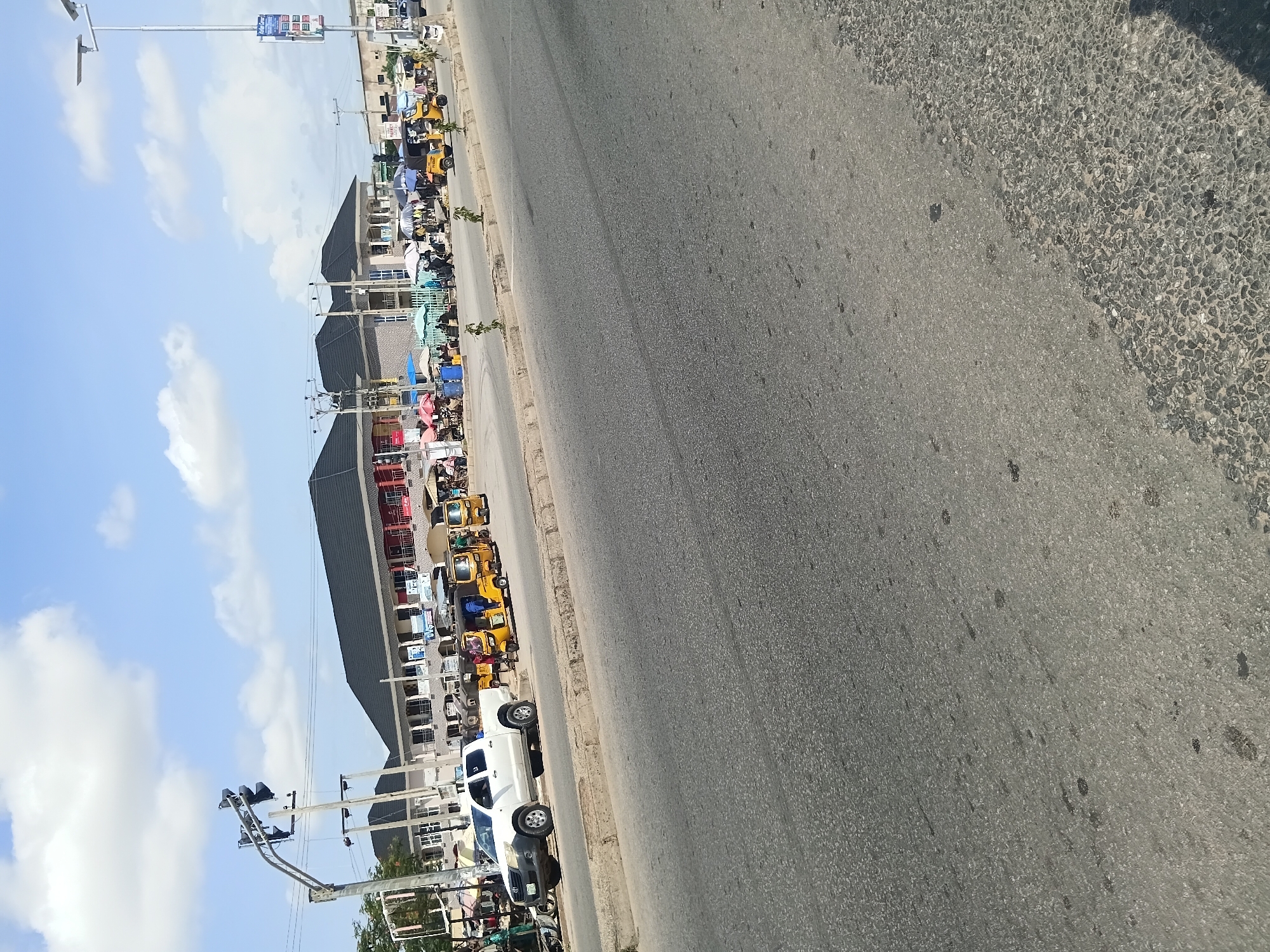 The image depicts a street scene in Jalingo, Nigeria, likely near a market or commercial area. In the foreground, a gray asphalt road occupies the majority of the frame, showing signs of wear and tear with some darker patches. The road is bordered by a concrete curb and a strip of rougher ground.
In the middle ground, several yellow auto-rickshaws, commonly known as 'keke napep' in Nigeria, are parked or moving. A white pickup truck is also visible on the side of the road. Behind the vehicles, a row of buildings with dark, sloped roofs lines the street. These buildings appear to be shops or commercial establishments, with awnings and some signs visible. Several individuals are present near these buildings, some appearing to be vendors or shoppers, with umbrellas providing shade.
The background reveals a clear blue sky with scattered white clouds. A traffic light structure stands on the left side of the image, and a lamppost with solar panels is visible on the right. The overall impression is a busy, everyday scene in an urban environment.
